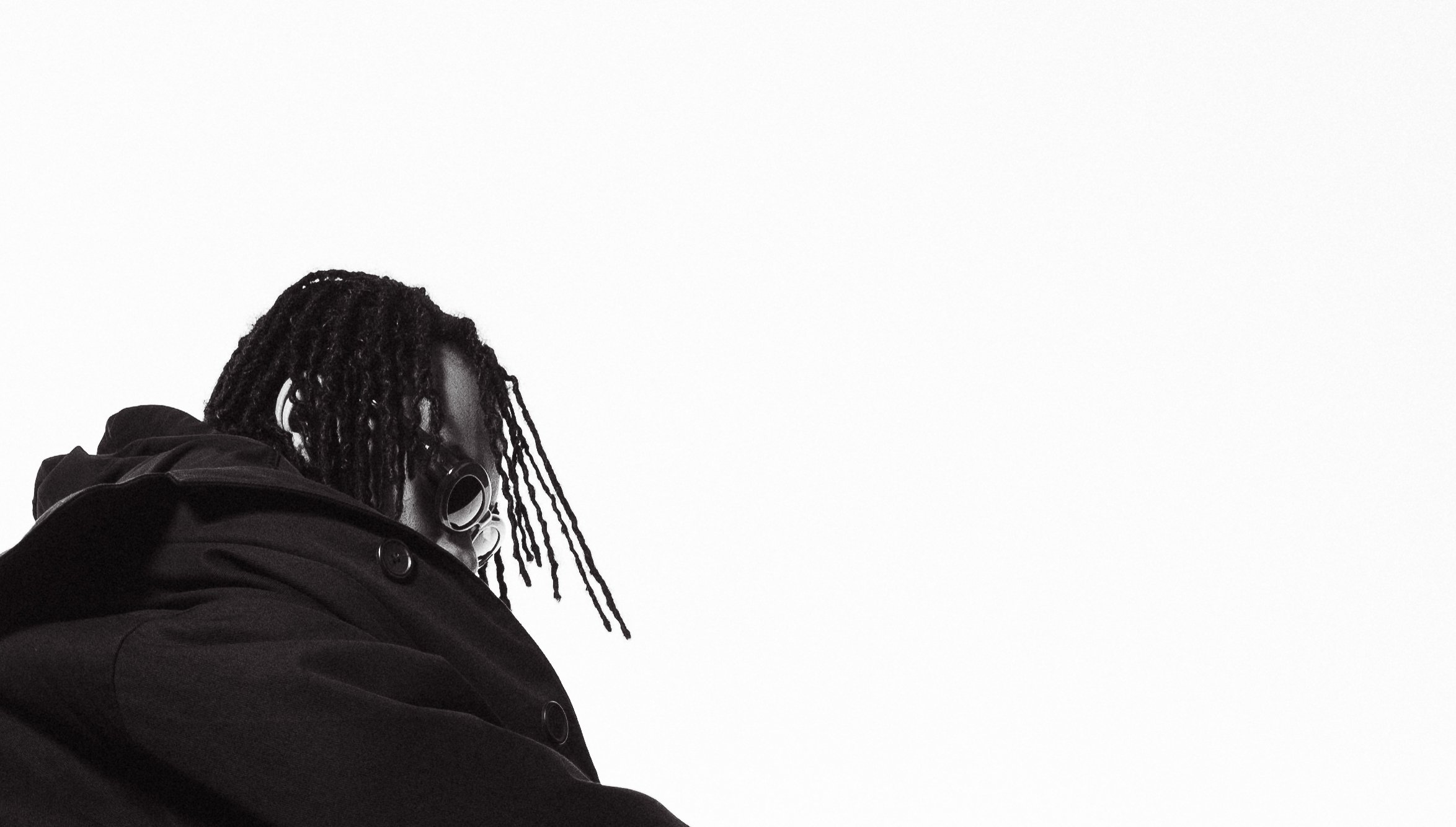 A person with long dreadlocks wearing headphones and a dark jacket, looking downward, against a plain white background.