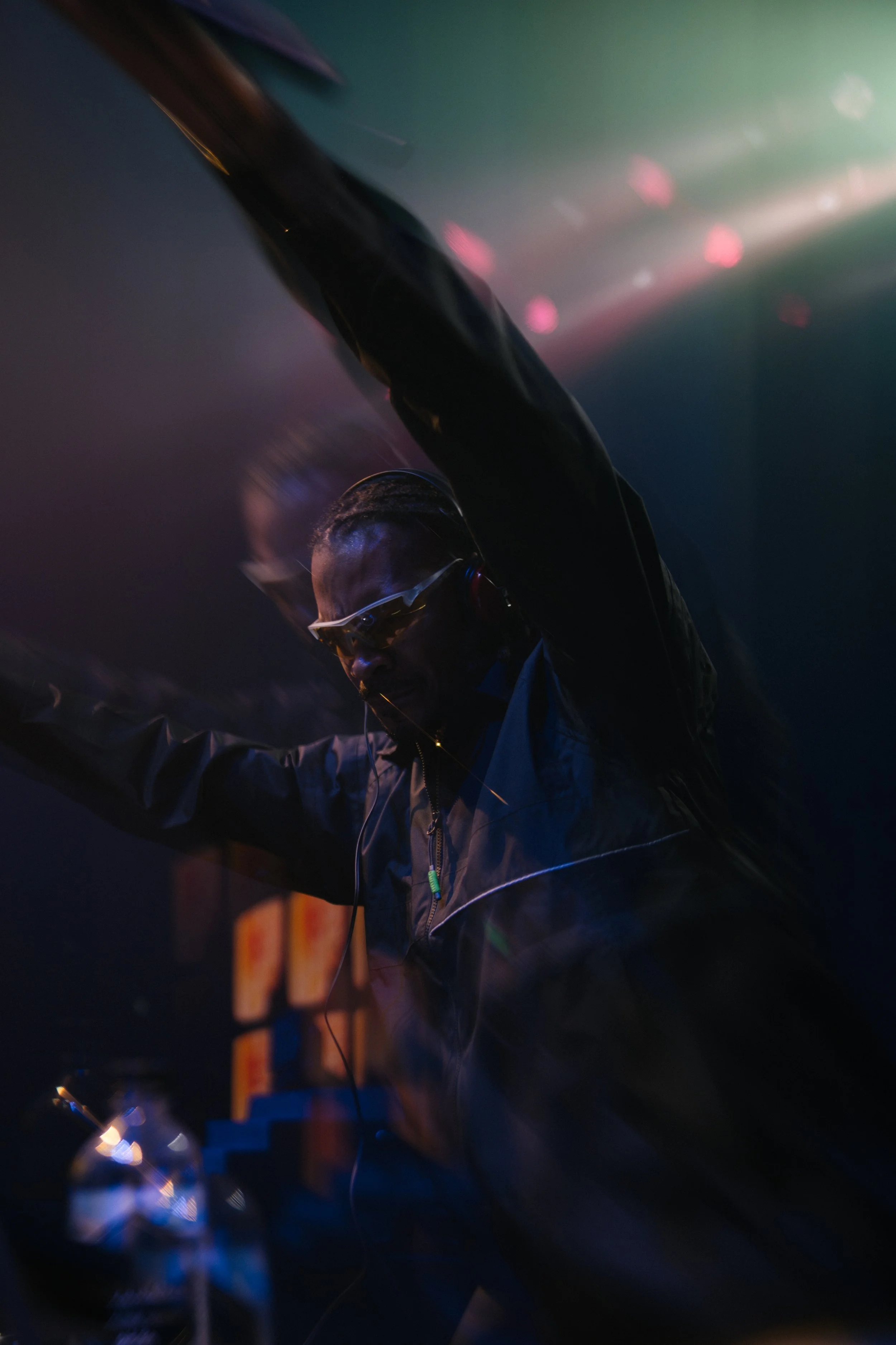 A DJ wearing glasses and headphones is mixing music in a dark, colorful club with pink and green lights.