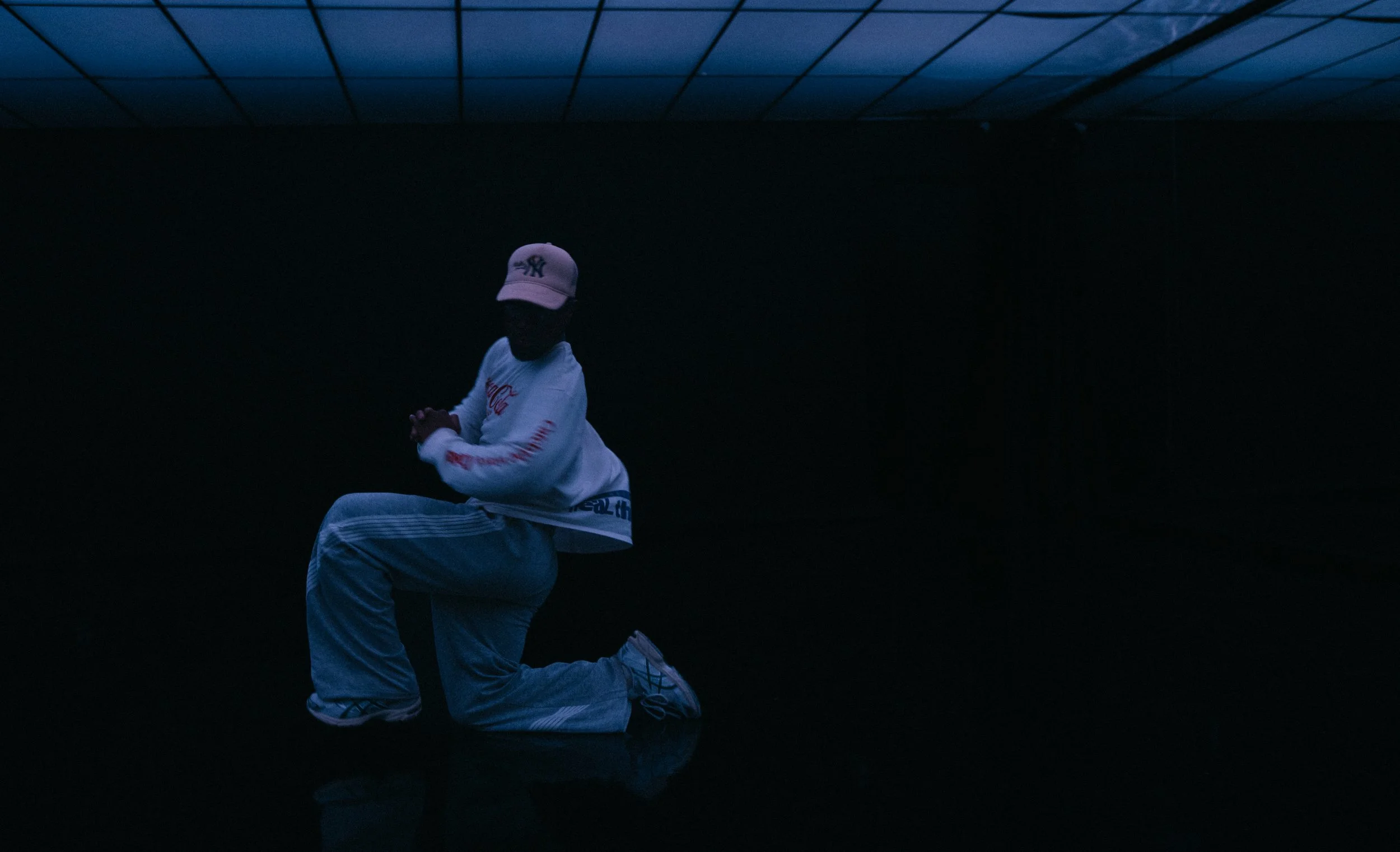 A person kneeling on the ground in a dark environment, wearing a white hoodie, light-colored pants, a pink cap, and sneakers.
