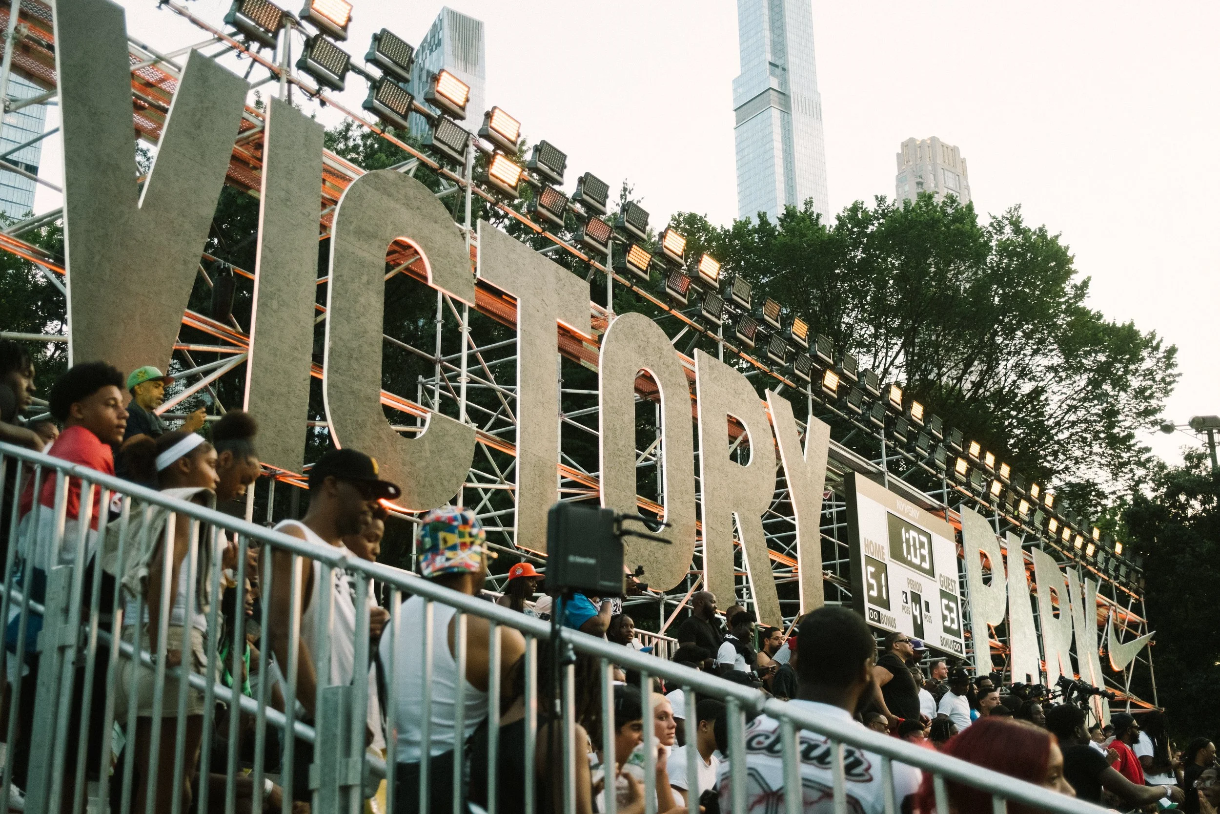 People gathered at an outdoor sports event with a large sign reading 'NY CARRY' and a digital scoreboard showing a score of 5-53.