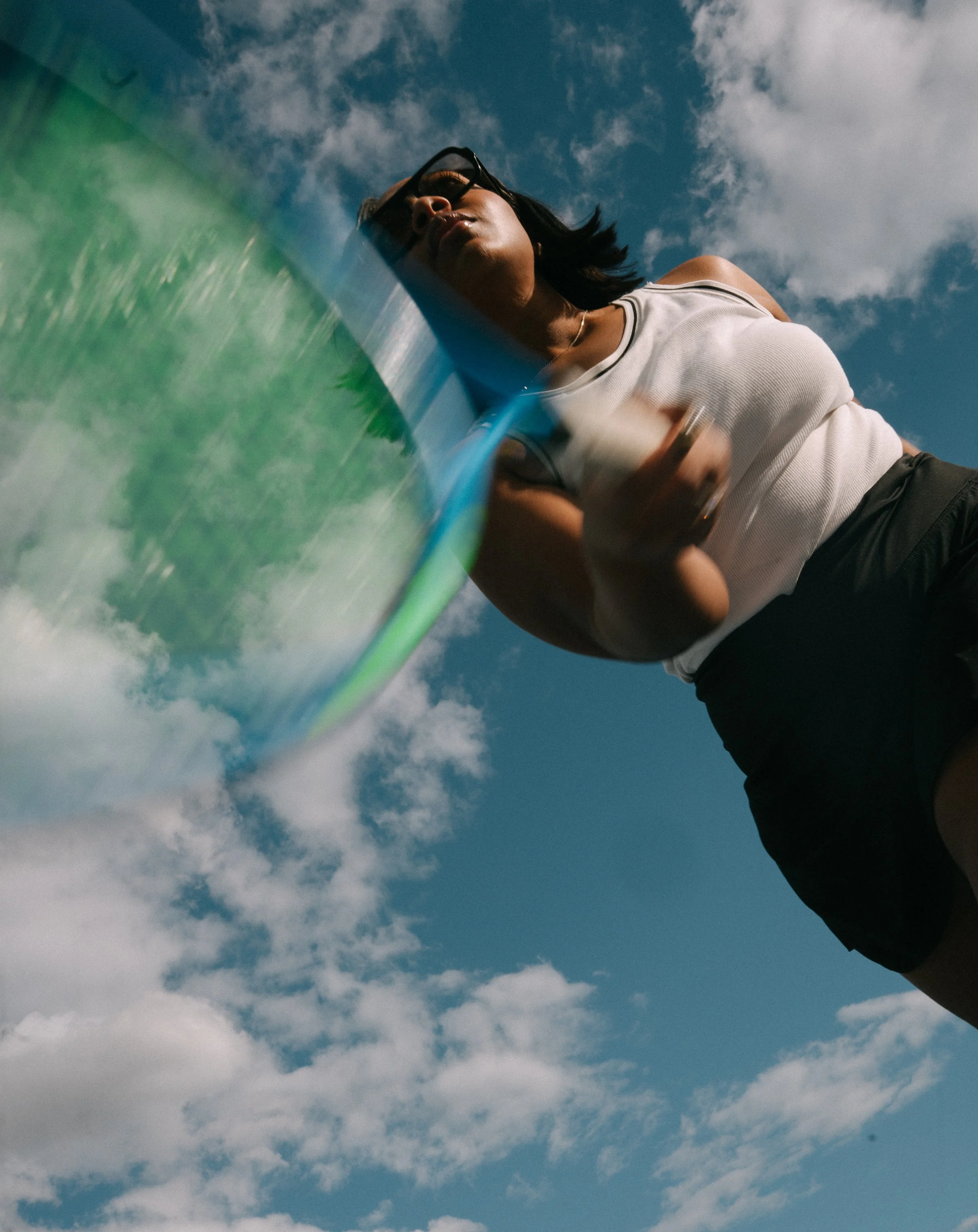Low-angle shot of a woman with sunglasses looking into the camera, holding a frisbee, against a partly cloudy sky.