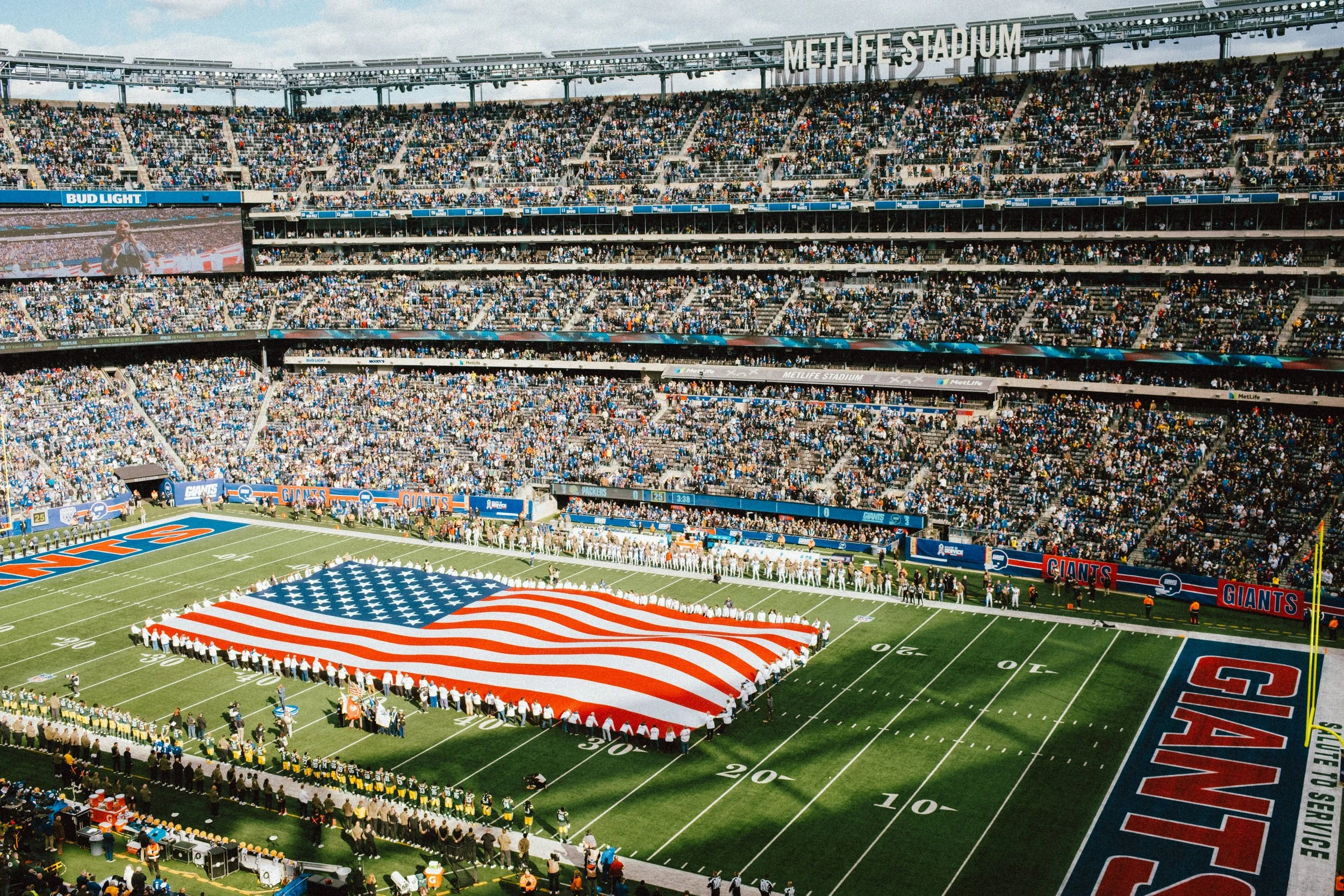 Crowd filling MetLife Stadium as players hold a large American flag on the field during a game, with the field marked with Yankees branding.