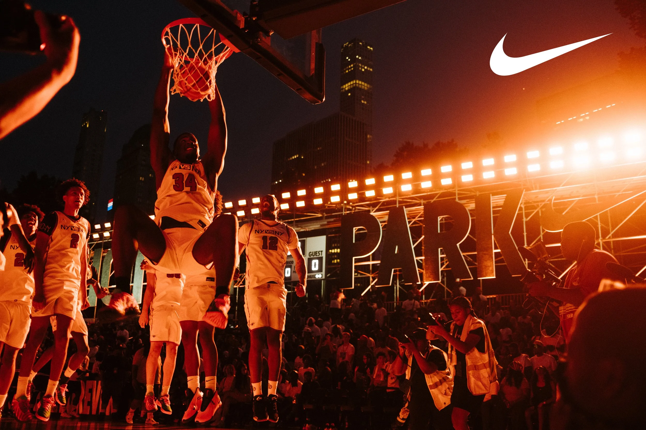 A basketball player wearing a jersey number 34 is dunking the ball, surrounded by teammates, during a game at night in an outdoor arena with city buildings in the background. Bright lights and a large sign spelling 'PARK' are visible, along with seve