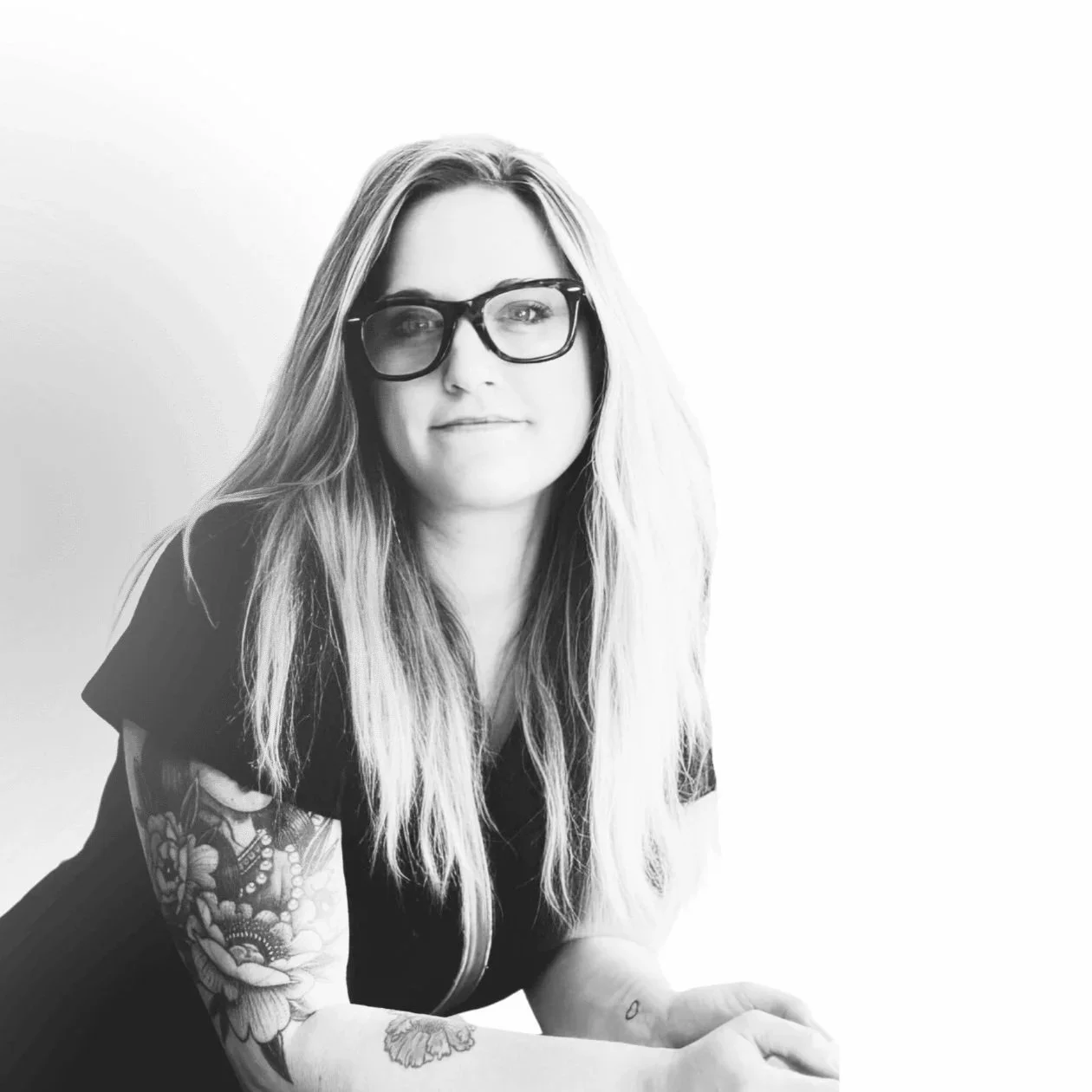 Black and white portrait of a woman with long hair wearing glasses, with tattoos on her arms, looking at the camera with a slight smile, against a plain white background.