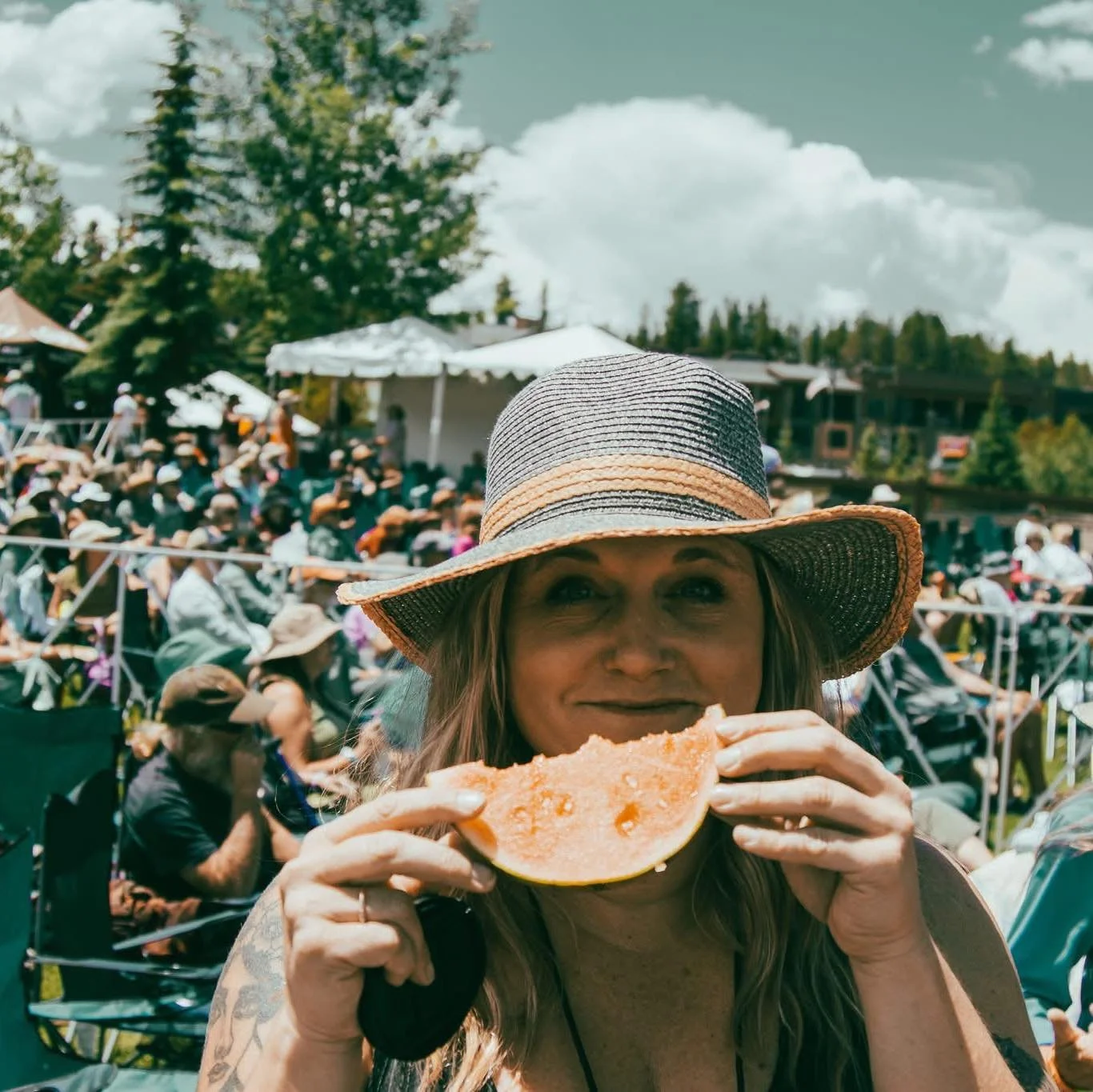 Woman wearing a large sun hat holding a slice of watermelon at an outdoor event with many seated people and trees in the background.