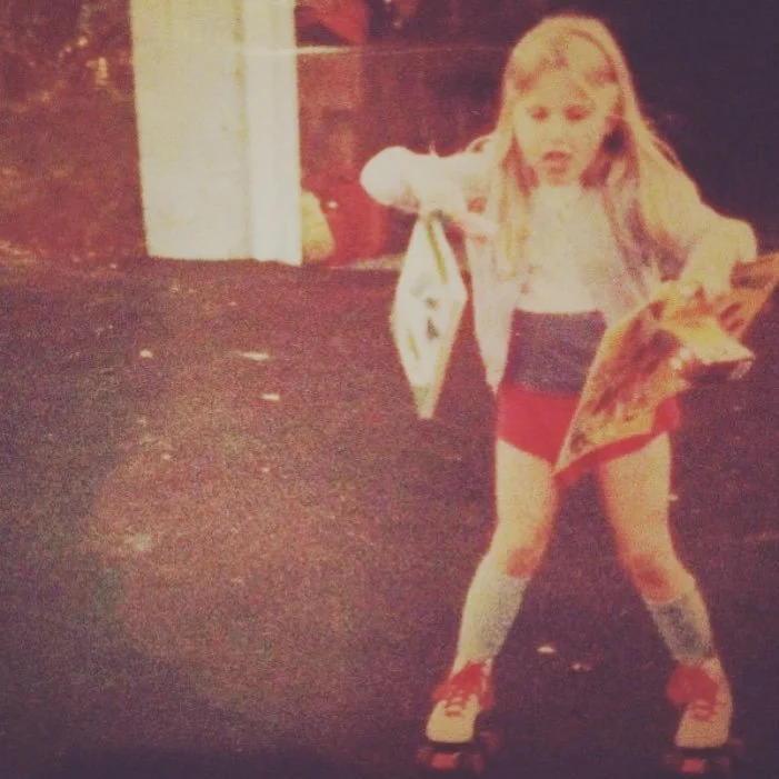 Young girl in roller skates holding a paper and a shopping bag, dressed in a white long-sleeve shirt and red shorts, skating outdoors at dusk.
