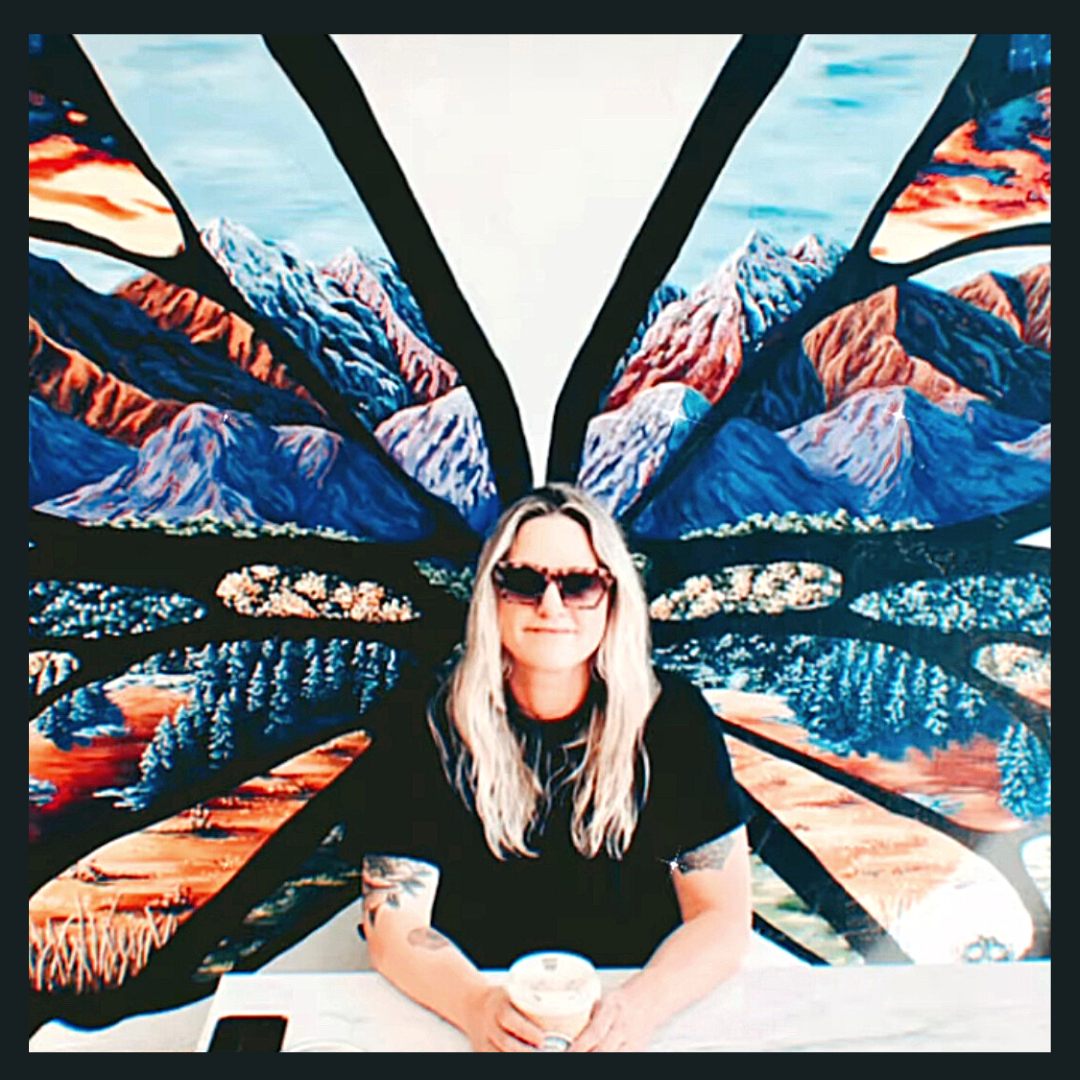 Woman with long blonde hair, wearing dark sunglasses and a black shirt, sitting at a table with a coffee drink, in front of a large, colorful butterfly mural featuring mountain and landscape scenes.