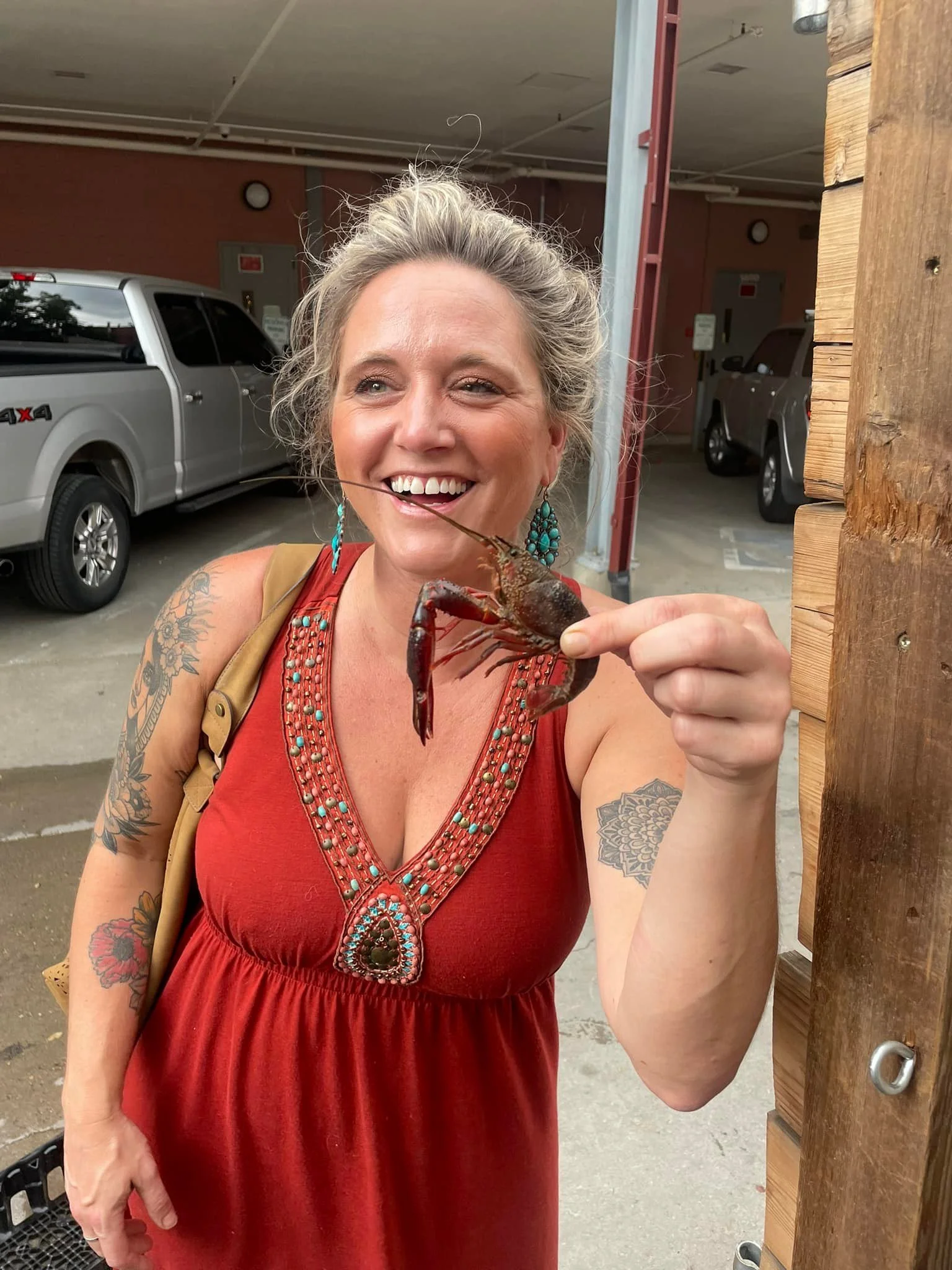 A woman with blonde hair, wearing a red dress with turquoise jewelry, is smiling and holding a large lobster.
