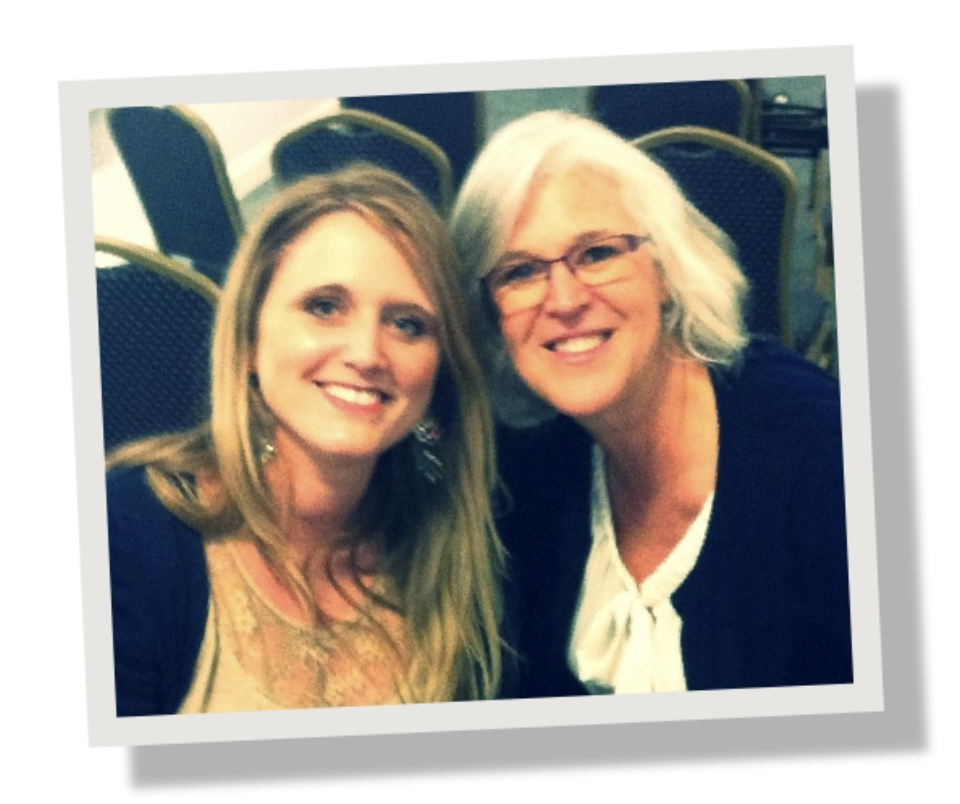 Two women smiling and sitting close together in a room with empty chairs in the background.