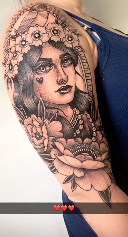 A black and gray tattoo of a woman with long hair, wearing a flower crown, jewelry, and an expressionless face, surrounded by flowers and leaves on her upper arm.