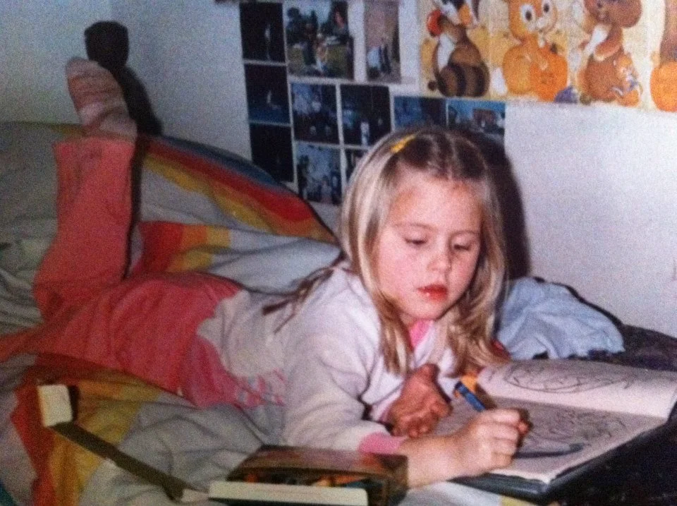 A young girl with blonde hair lying on her stomach on a bed, coloring in a book with crayons.