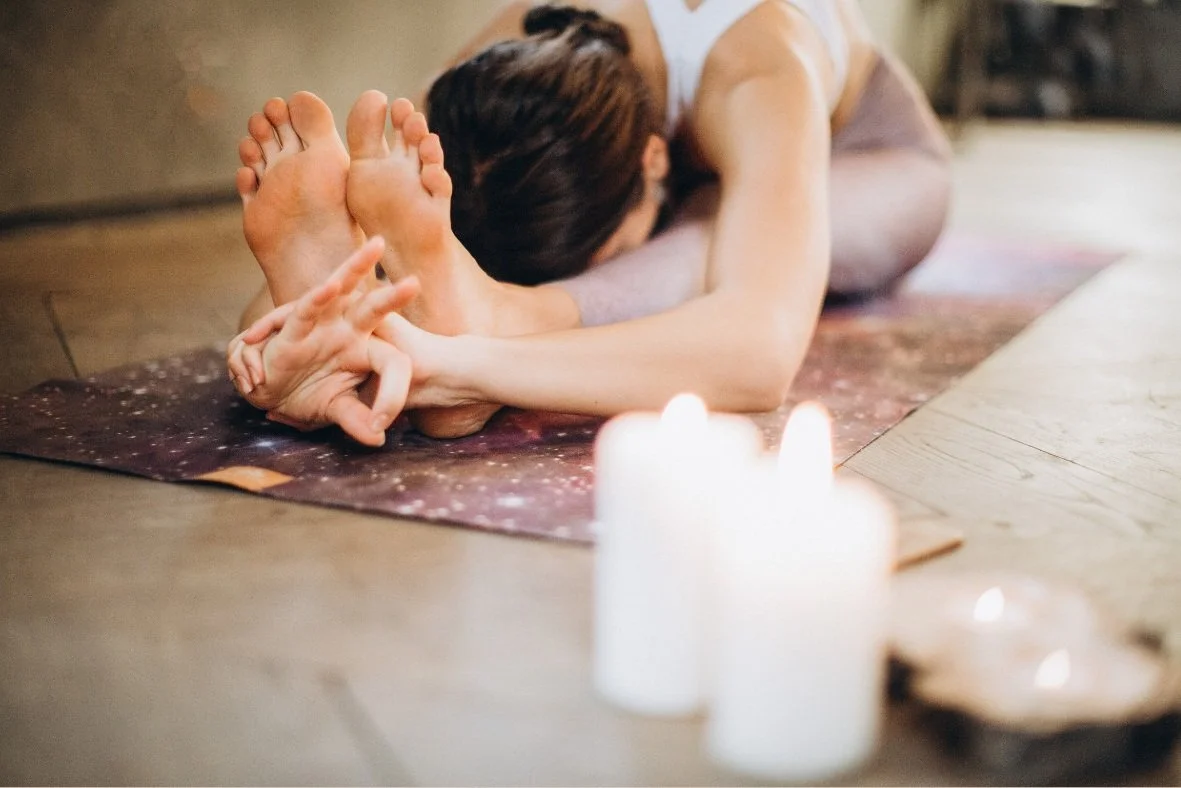 Person practicing yoga on a mat in a forward fold position with lit candles nearby.