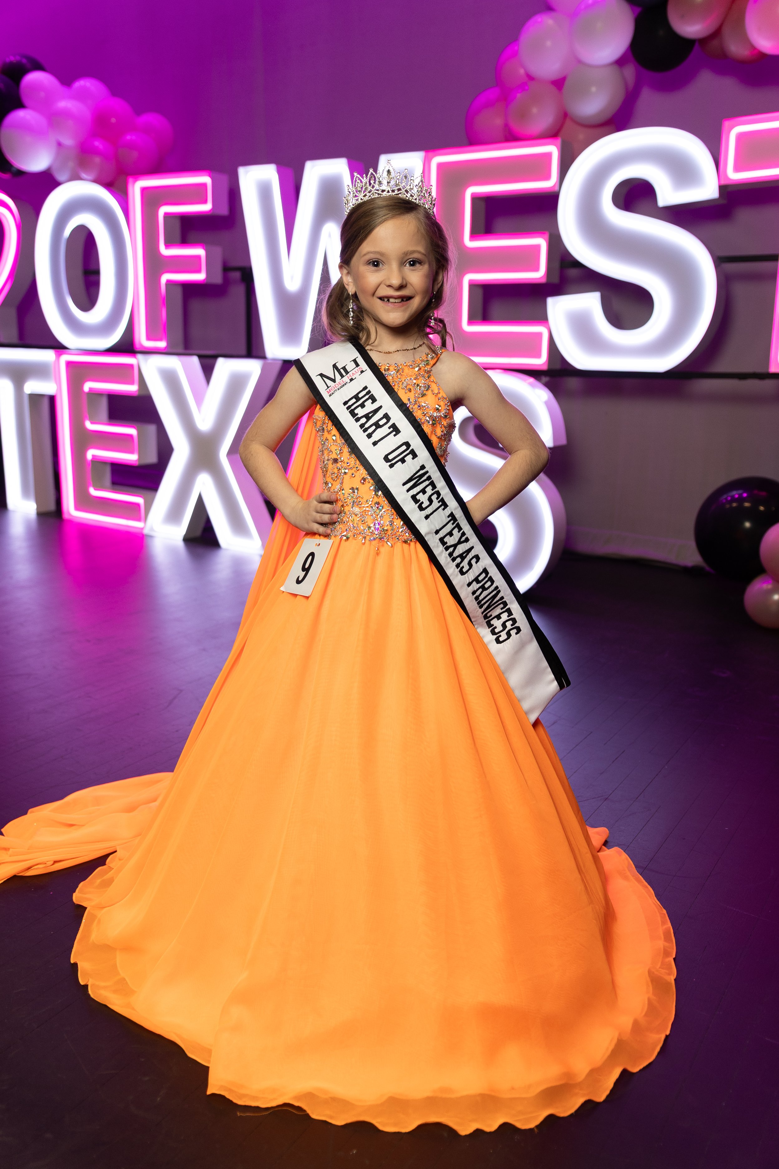 Young girl with long brown hair wearing a pink gown, crown, earrings, and a sash that reads 'Heart of West Texas Princess'.