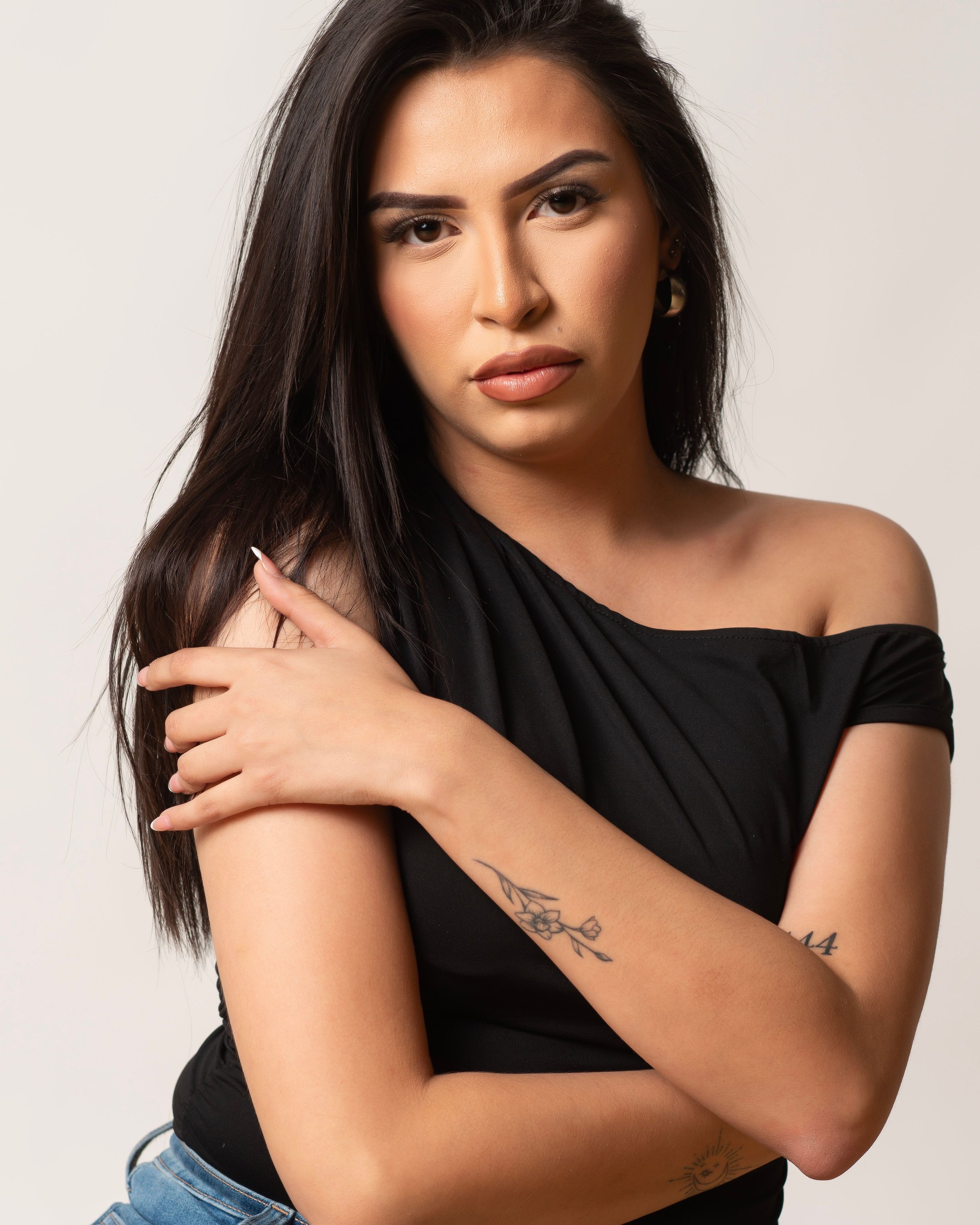 A woman with long dark hair and light skin looking directly at the camera, wearing a black off-shoulder top, with tattoos on her arm and side, and ear cuffs.