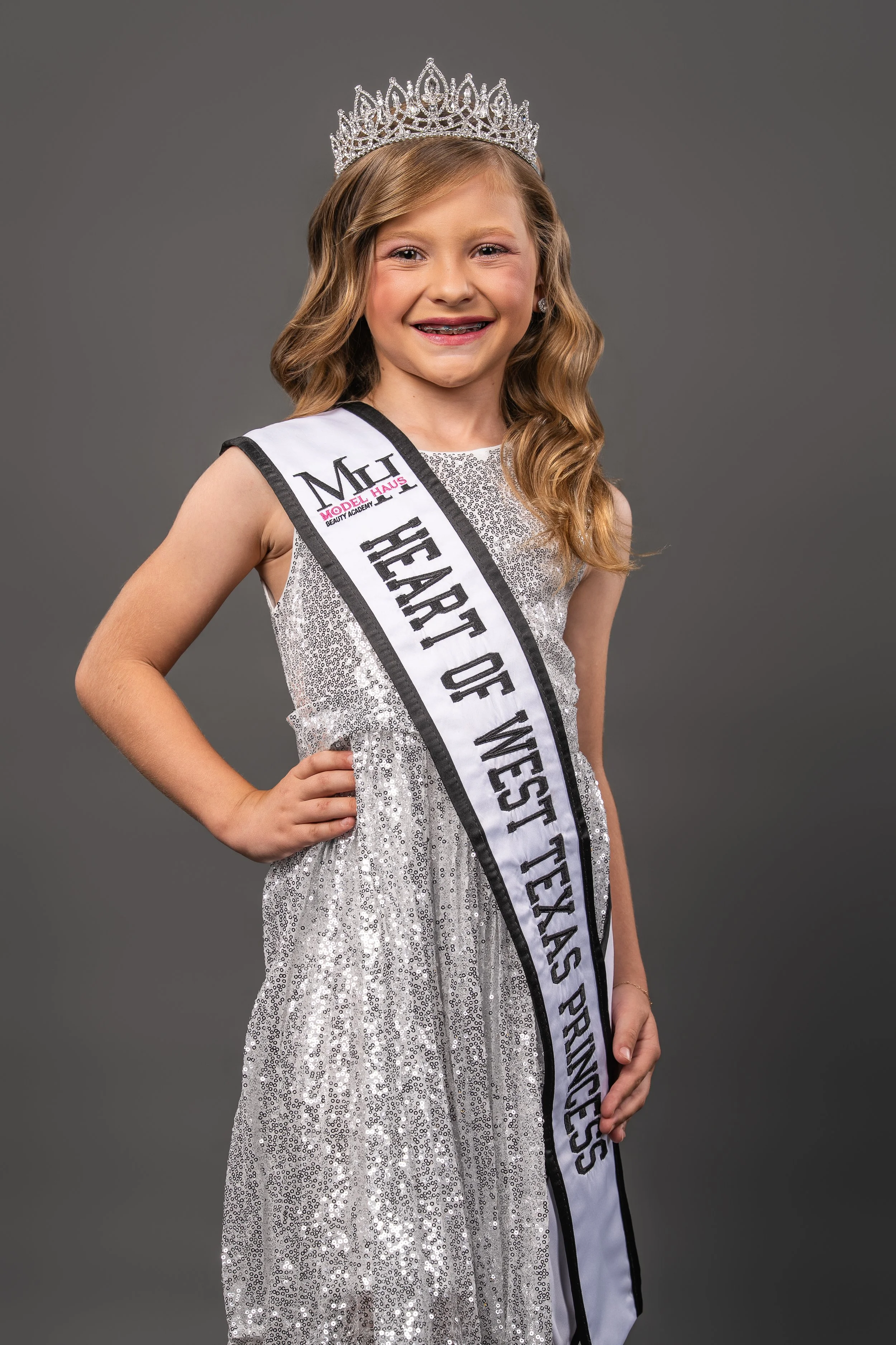 Young girl with long brown hair wearing a pink gown, crown, earrings, and a sash that reads 'Heart of West Texas Princess'.