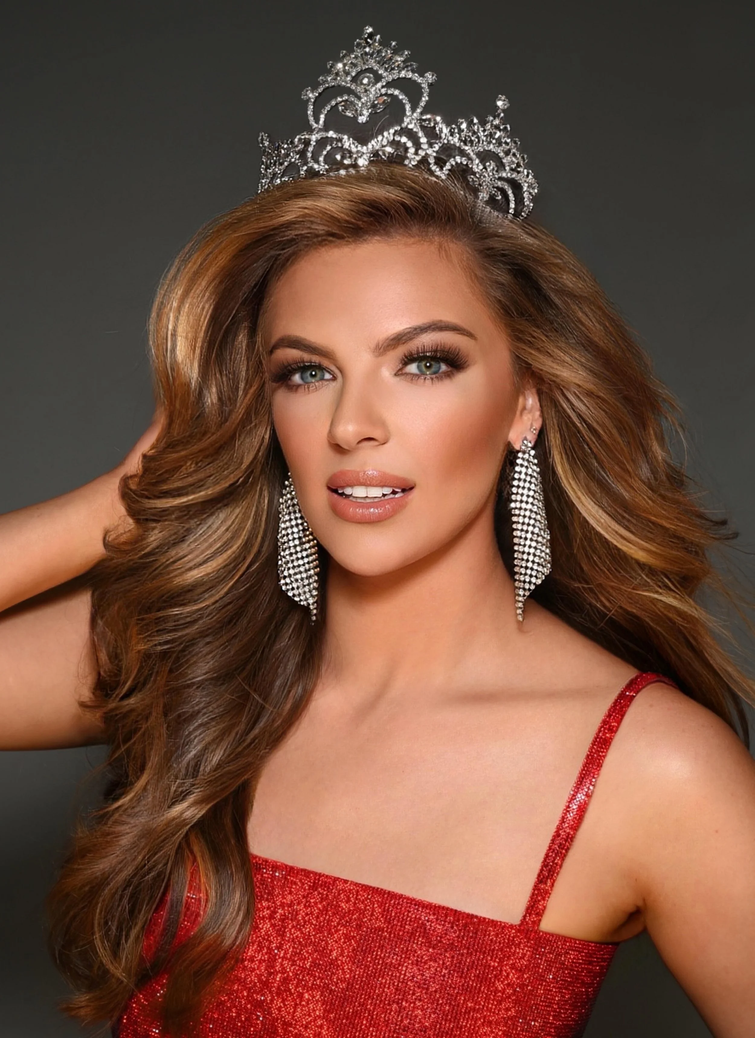 A woman with long, wavy, light brown hair wearing a silver tiara and large, dangling earrings. She is dressed in a red, sparkly dress with thin straps and is posing against a dark gray background.