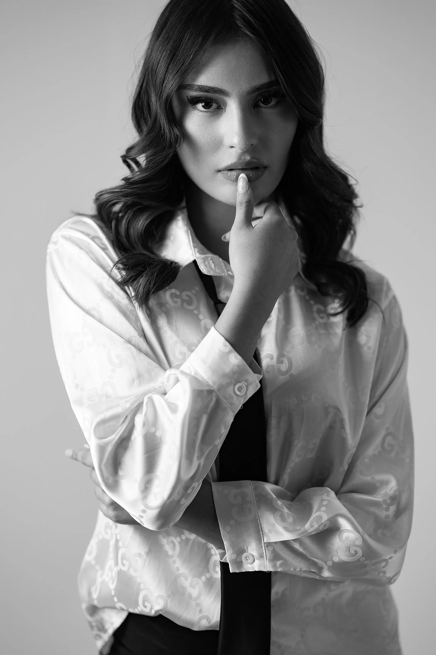 Black and white portrait of a woman with wavy hair, wearing a patterned satin shirt and a dark tie, looking directly at the camera with her finger on her lips.