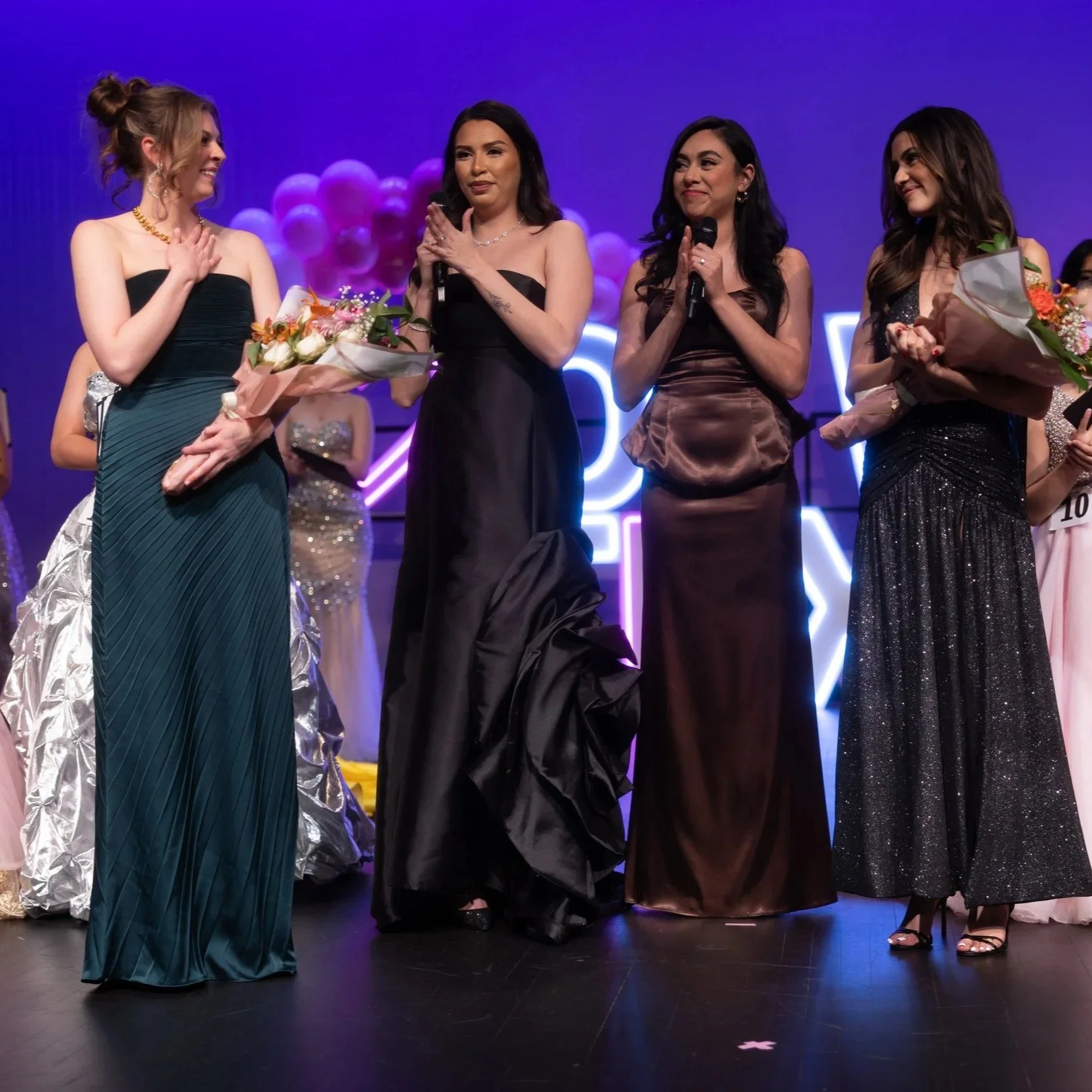 Four women in elegant evening gowns stand on stage during an event, with one woman holding a bouquet of flowers and the others holding microphones, colorful balloons in the background.