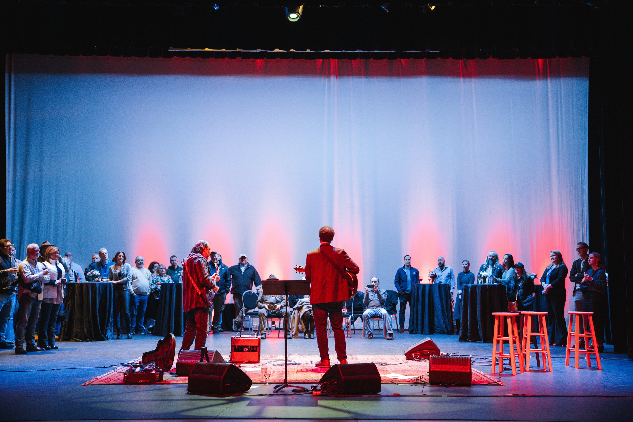 A musician with a guitar performing on stage with a large audience watching. The stage is lit with colorful lights and has a simple setup with stools and standing tables.