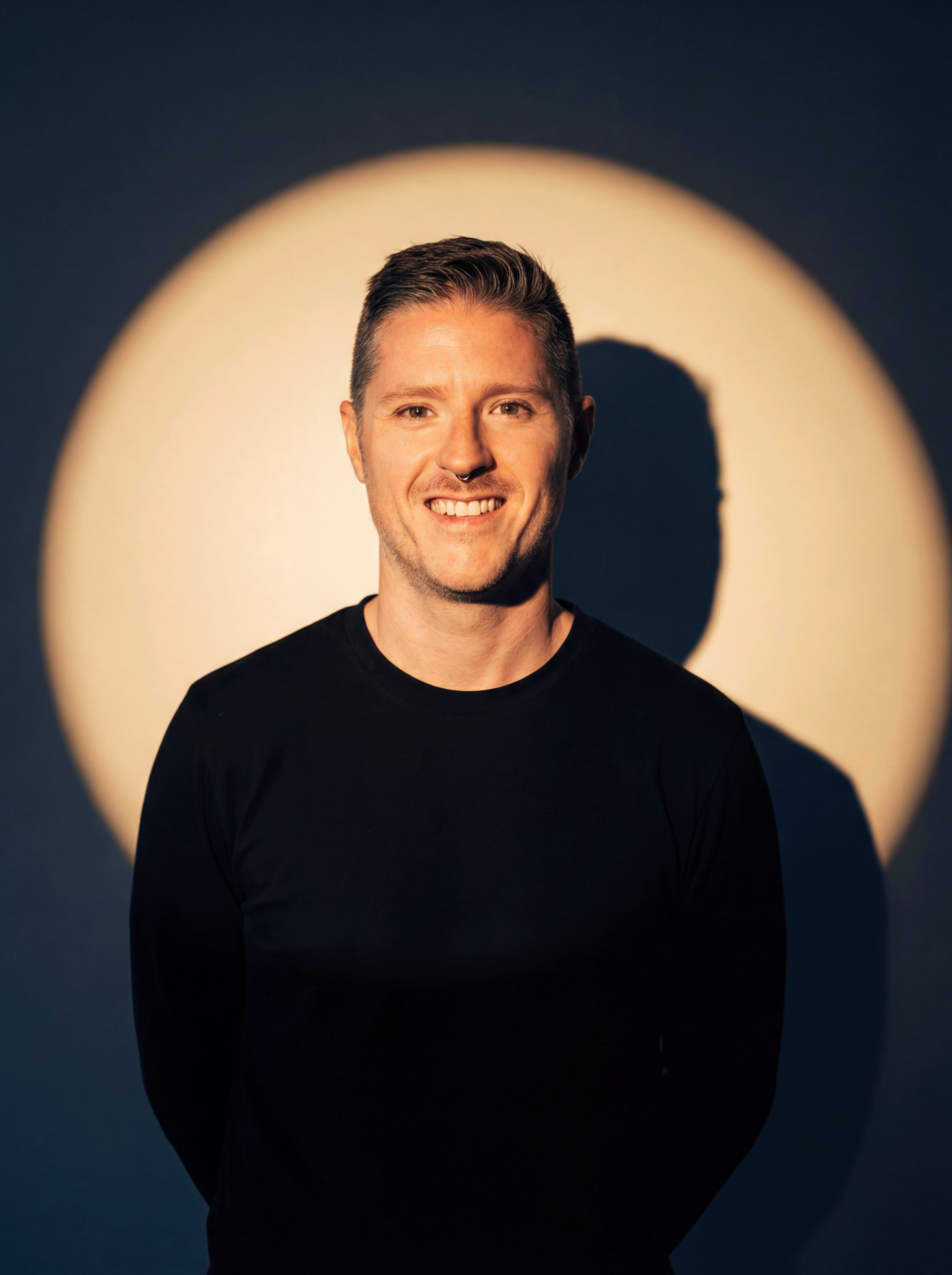 Portrait of a man with short brown hair and a small goatee, smiling, wearing a black shirt, standing in front of a dark background with a large circular spot of light behind him.