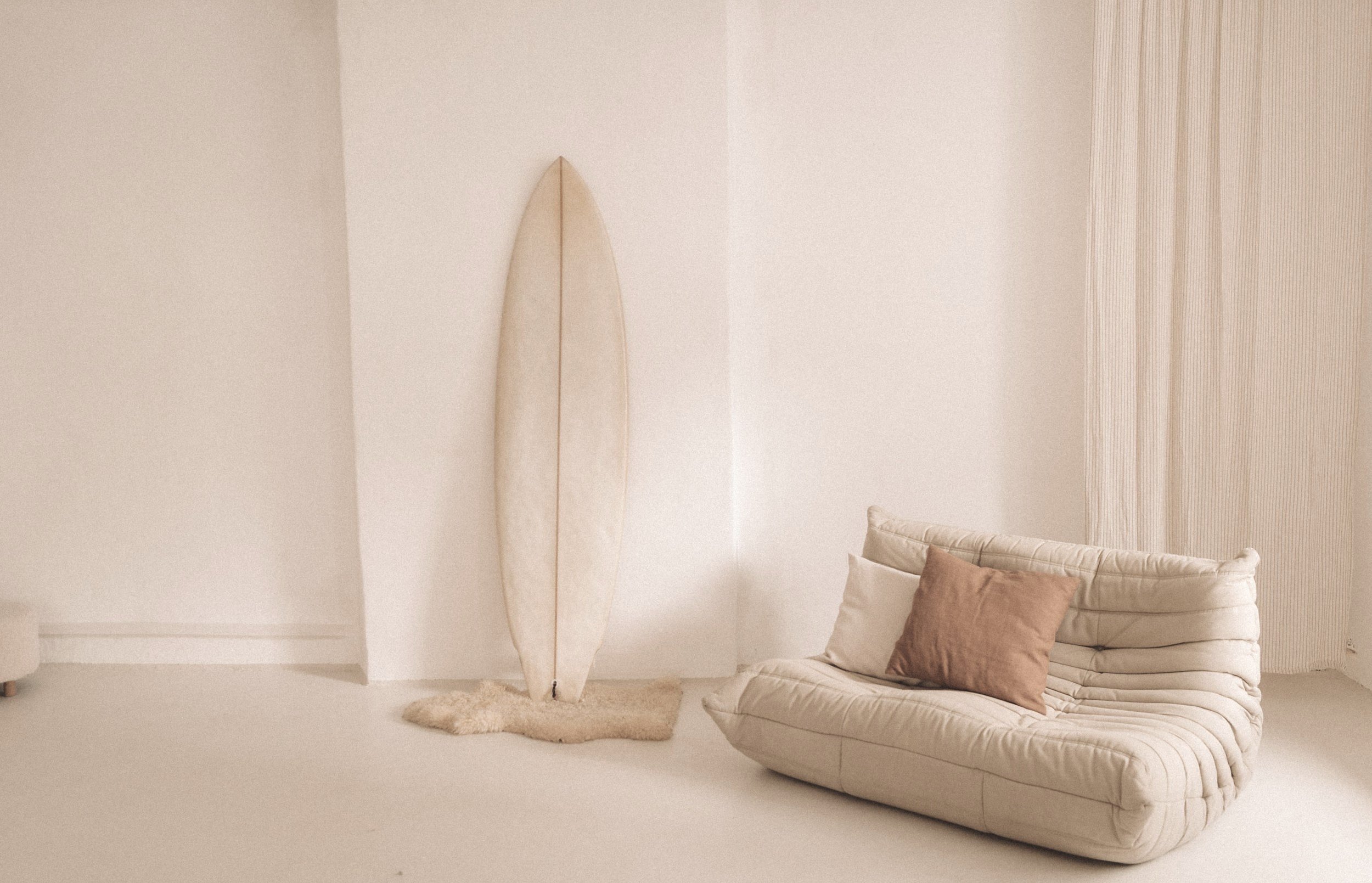 Minimalist room with a beige surfboard leaning against a wall, a beige cushioned sofa with pillows, and soft lighting.