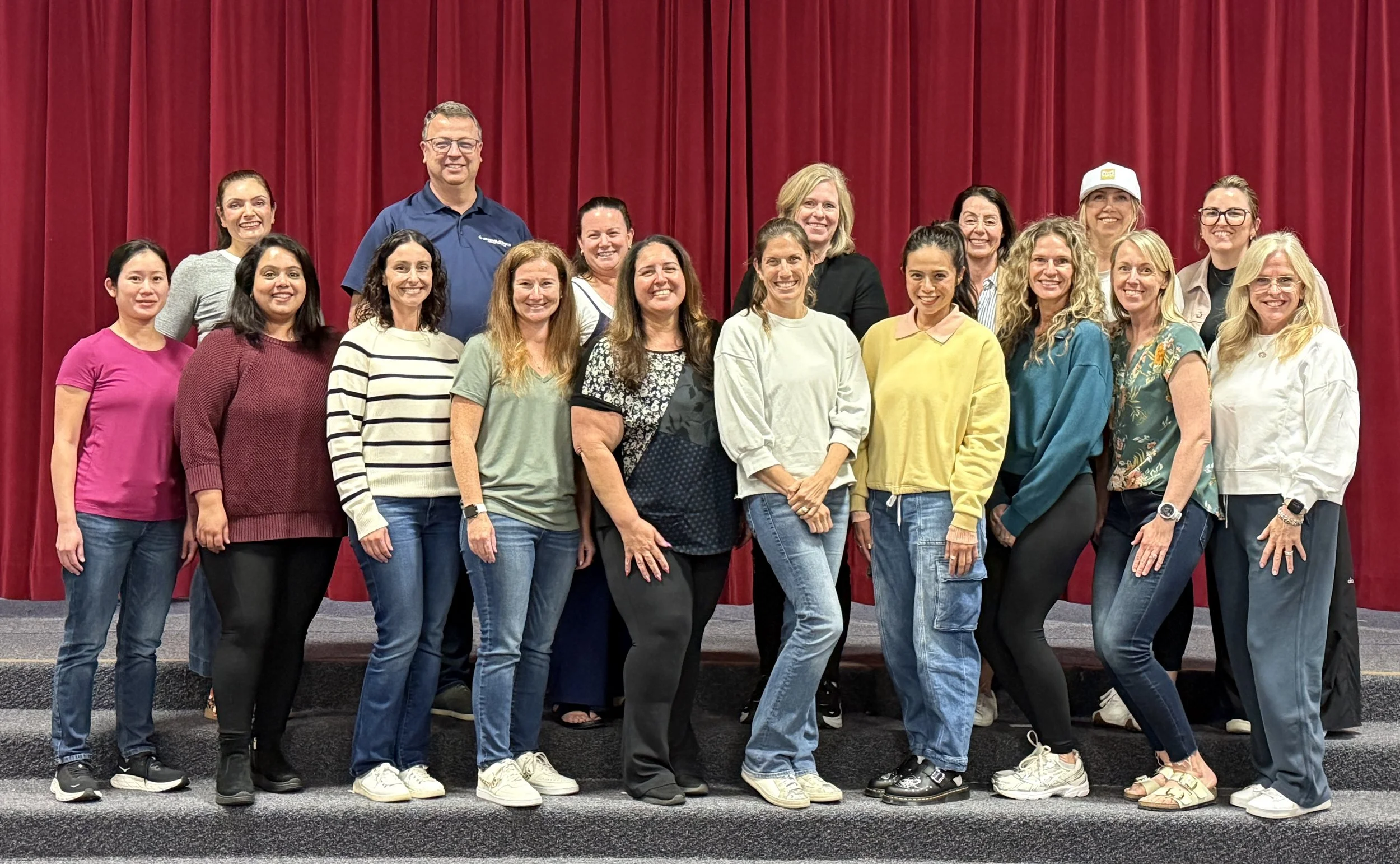 A group of fifteen women and one man standing on a stage in front of a red curtain, smiling at the camera, with some women wearing casual clothes and one woman wearing a white cap.
