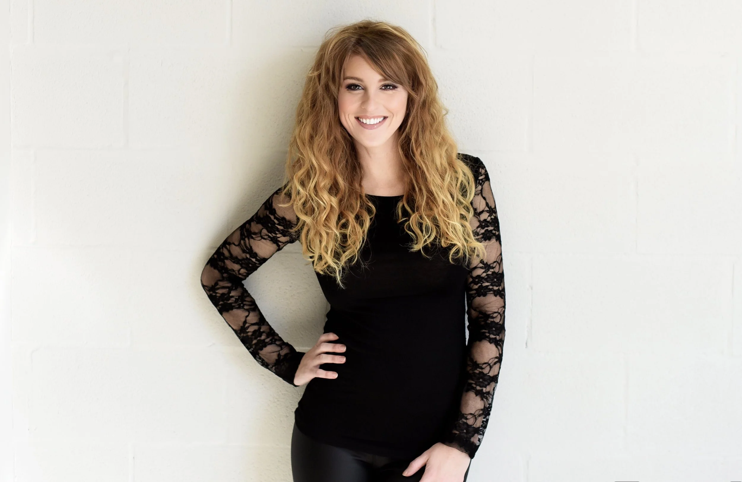 A woman with long curly red hair smiling, wearing a black dress with lace sleeves, standing against a white brick wall with her right hand on her hip.