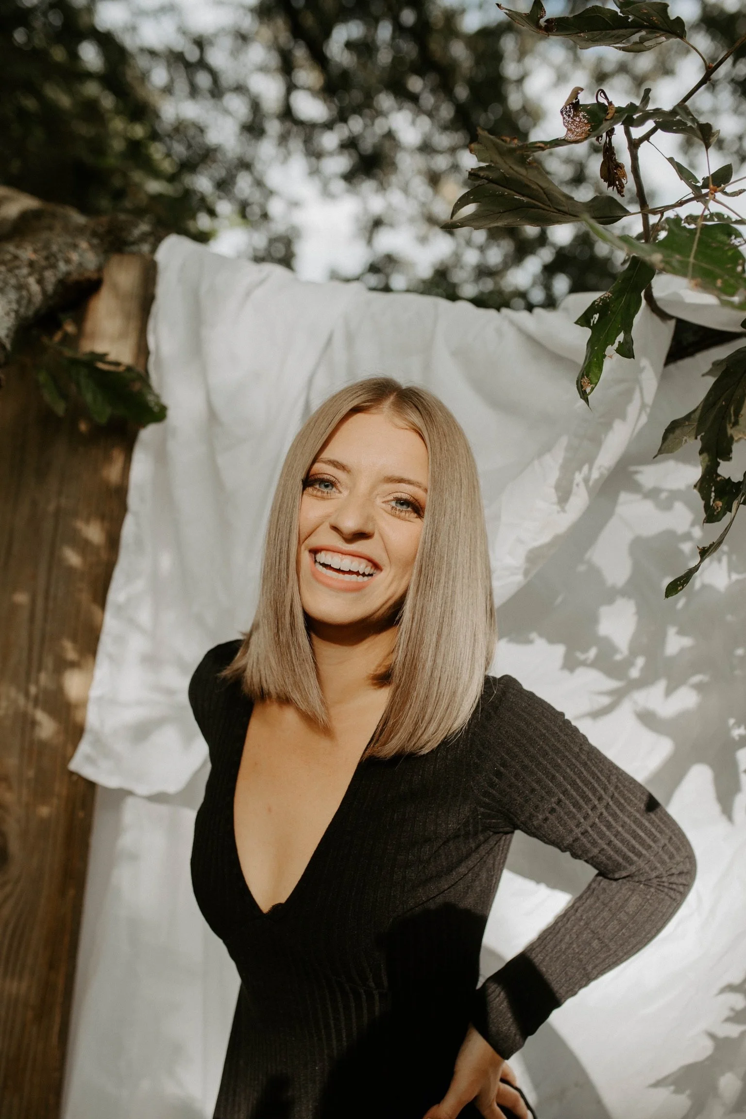 A woman with blond hair smiling, wearing a black dress, standing in front of a white sheet backdrop with tree branches and leaves overhead.