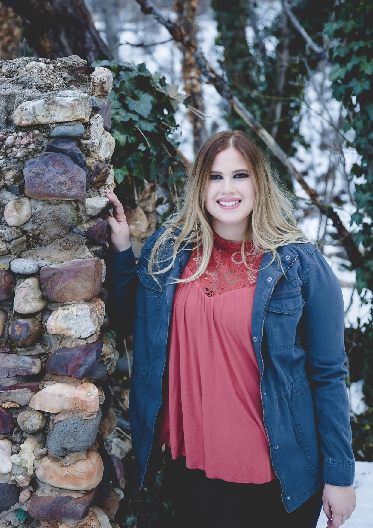 Girl with blonde hair wearing a blue jacket and red lace top standing outdoors near a stone wall.