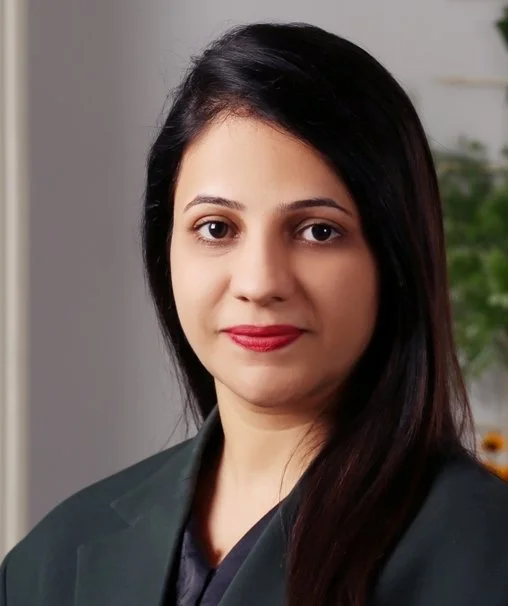 Professional woman with long dark hair, wearing a dark blazer, posing in front of a light-colored background with a plant in the background.