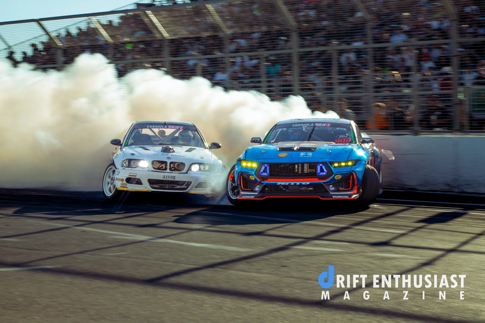 Two race cars drifting on a track, with a crowd watching from the stands in the background and smoke billowing behind them, during a Formula Drift event.
