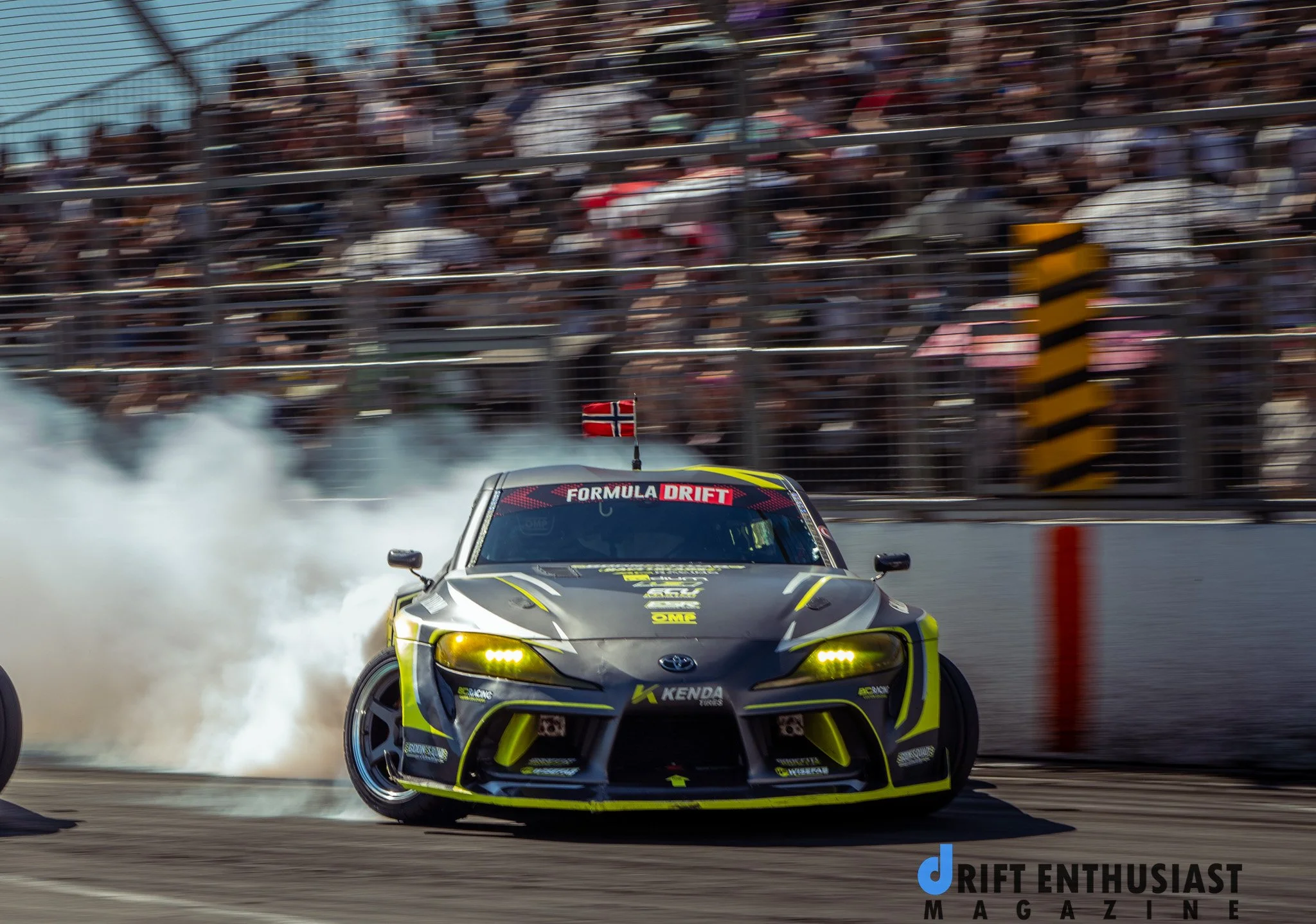 A black and yellow drift car with a Norwegian flag on top, drifting on a race track while smoke billows from the tires, with a crowd of spectators behind a fence.