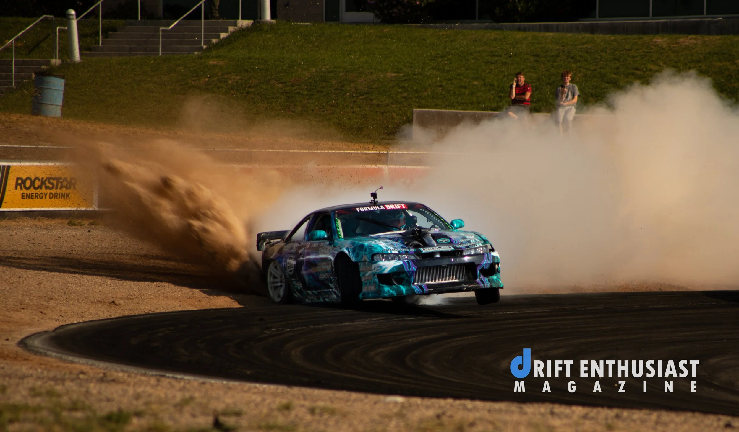 A drift car on a dirt racetrack kicking up dust as it rounds a turn, with two spectators watching from the hillside in the background. The car has a vibrant, patterned paint job and is marked with 'Formula Drift' and the 'Drift Enthusiast Magazine' logo.