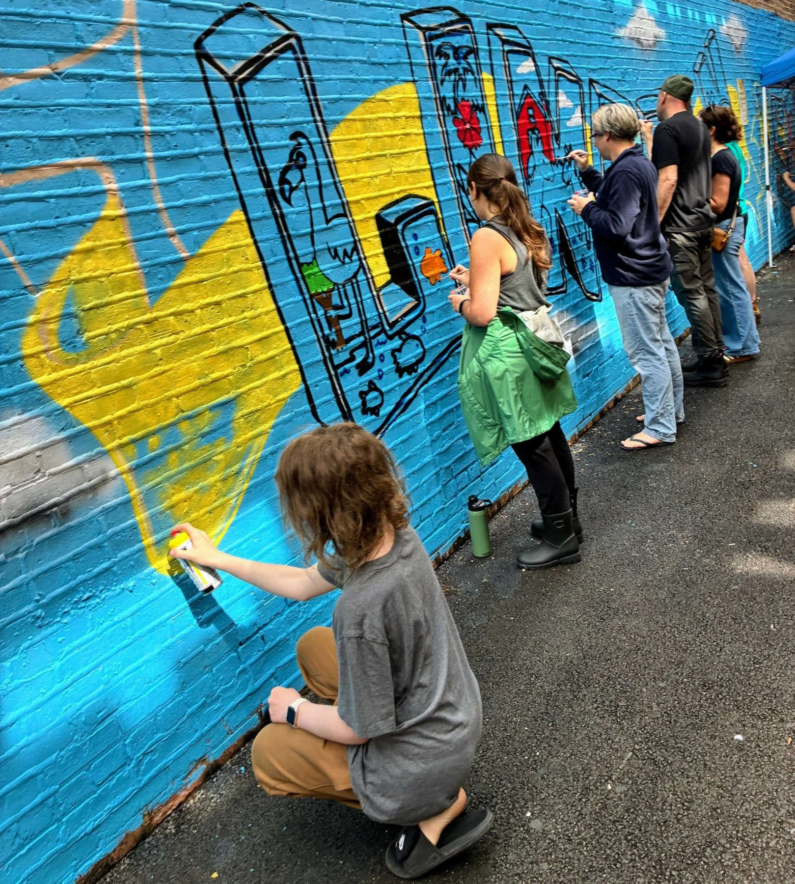 Community volunteers working together to paint colorful graffiti-style lettering and animal illustrations on a blue brick wall during the Lincoln Square Mural Project 2026 in Chicago.
