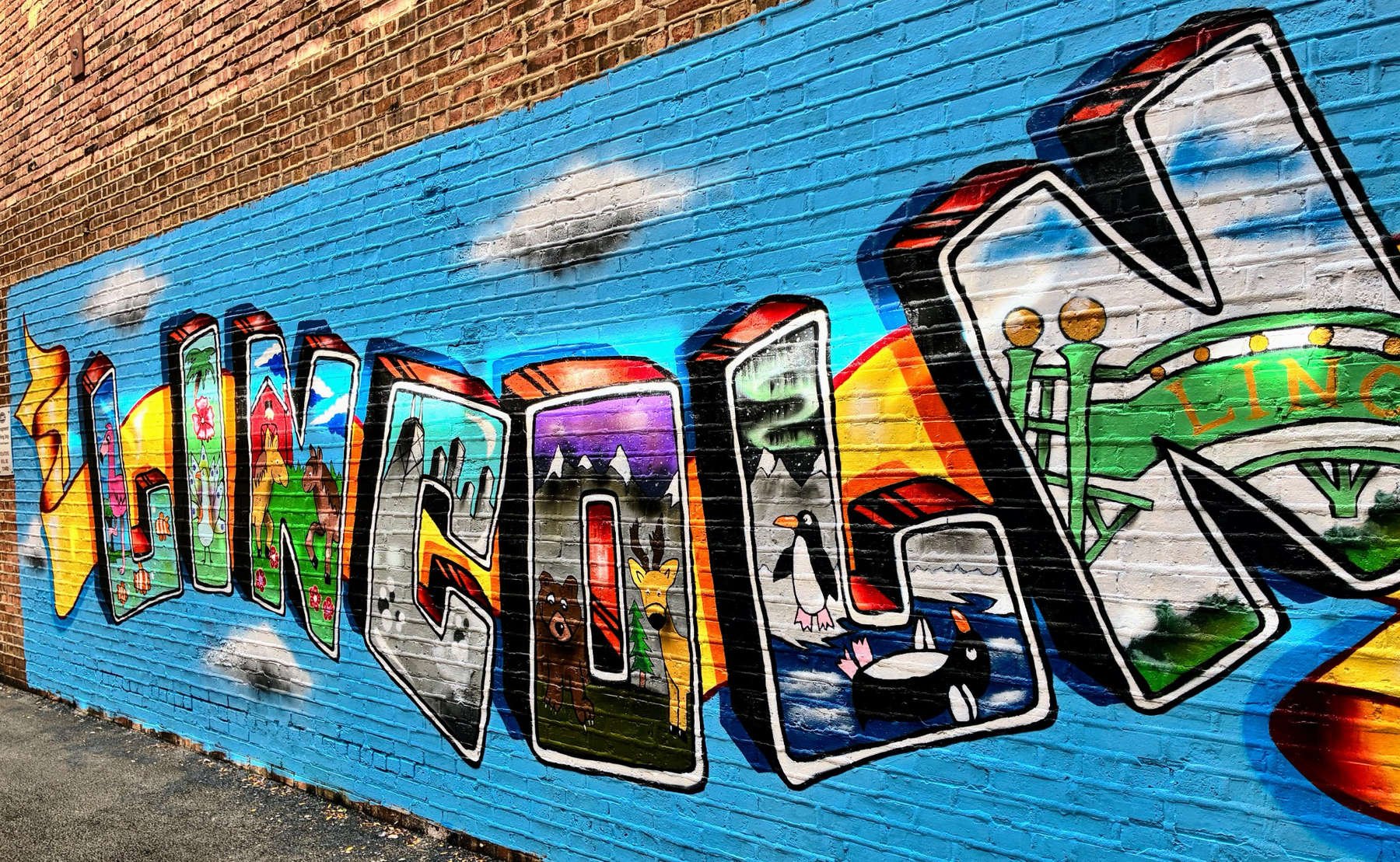 Completed LINCOLN lettering in the Lincoln Square community mural in Chicago, featuring colorful animal illustrations and landscapes within three-dimensional graffiti-style letters on a bright blue brick wall.