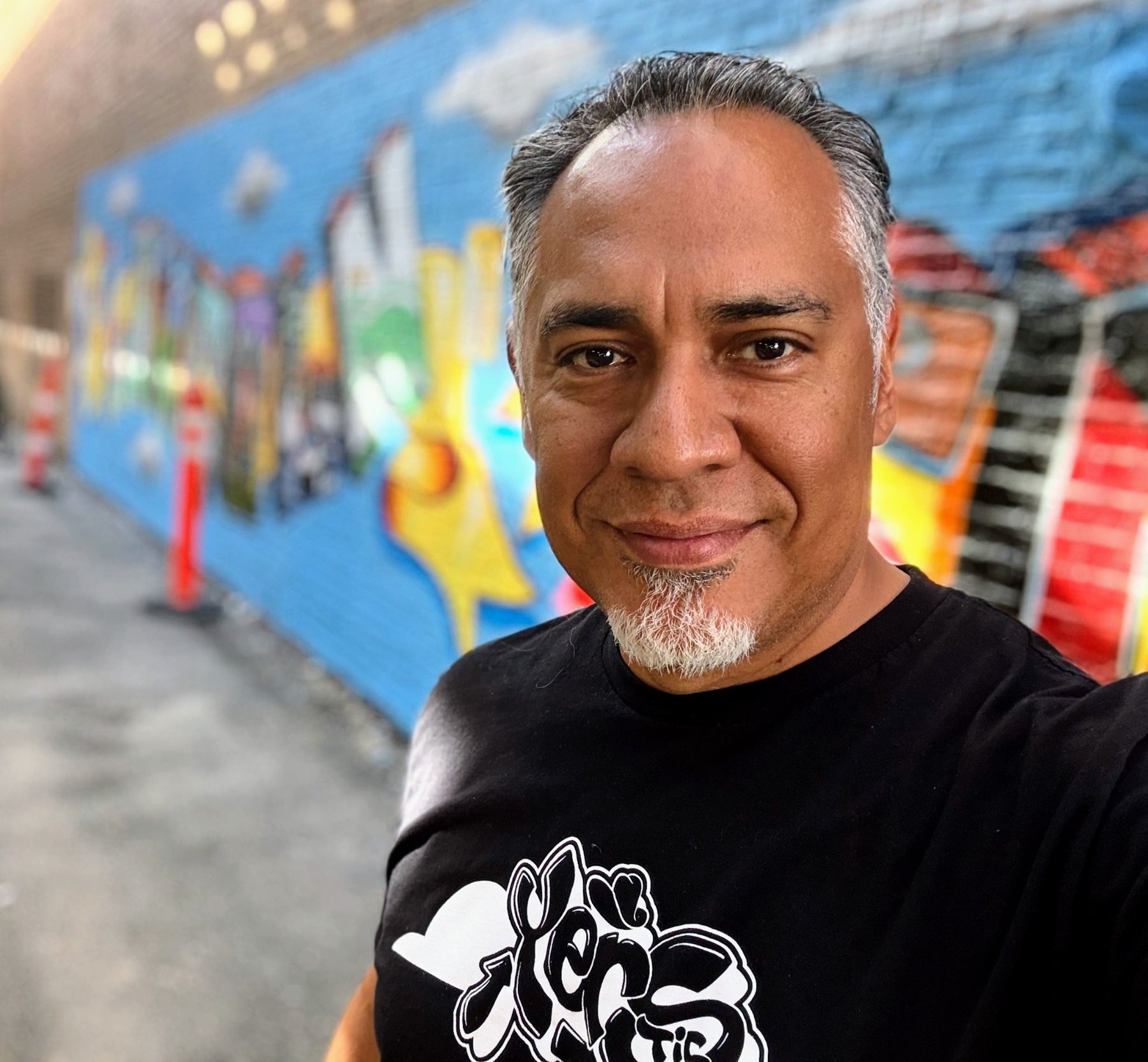 Chicago graffiti artist Nino “Tsel One/Tsel1” Rodriguez, lead artist of the Lincoln Square Mural Project 2026, standing in front of the colorful community mural beneath the CTA Brown Line.