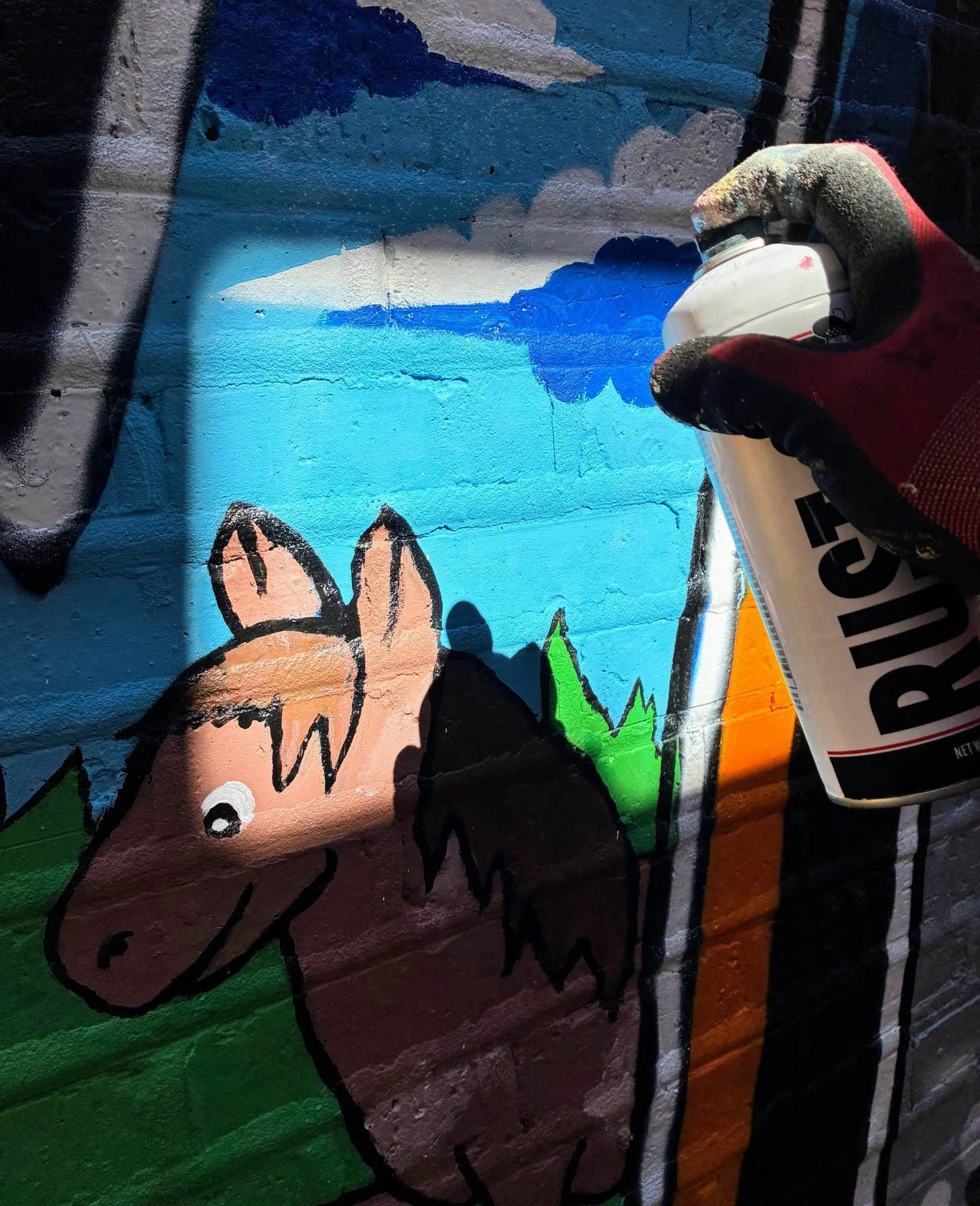 Close-up of a gloved hand using a spray-paint can to add colorful details to the Lincoln Square community mural on a blue brick wall in Chicago.