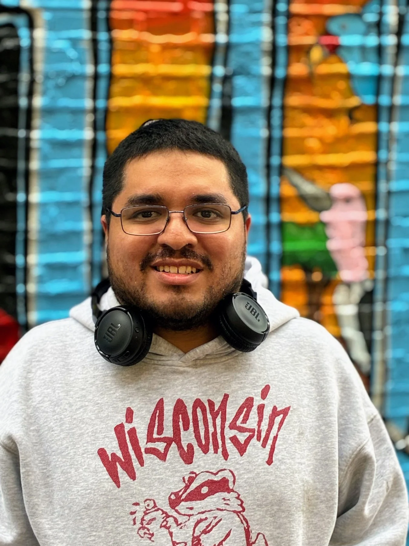 Chicago artist Mario Rodriguez standing in front of the colorful Lincoln Square community mural, a public art project supported by a DCASE grant awarded to Mario.