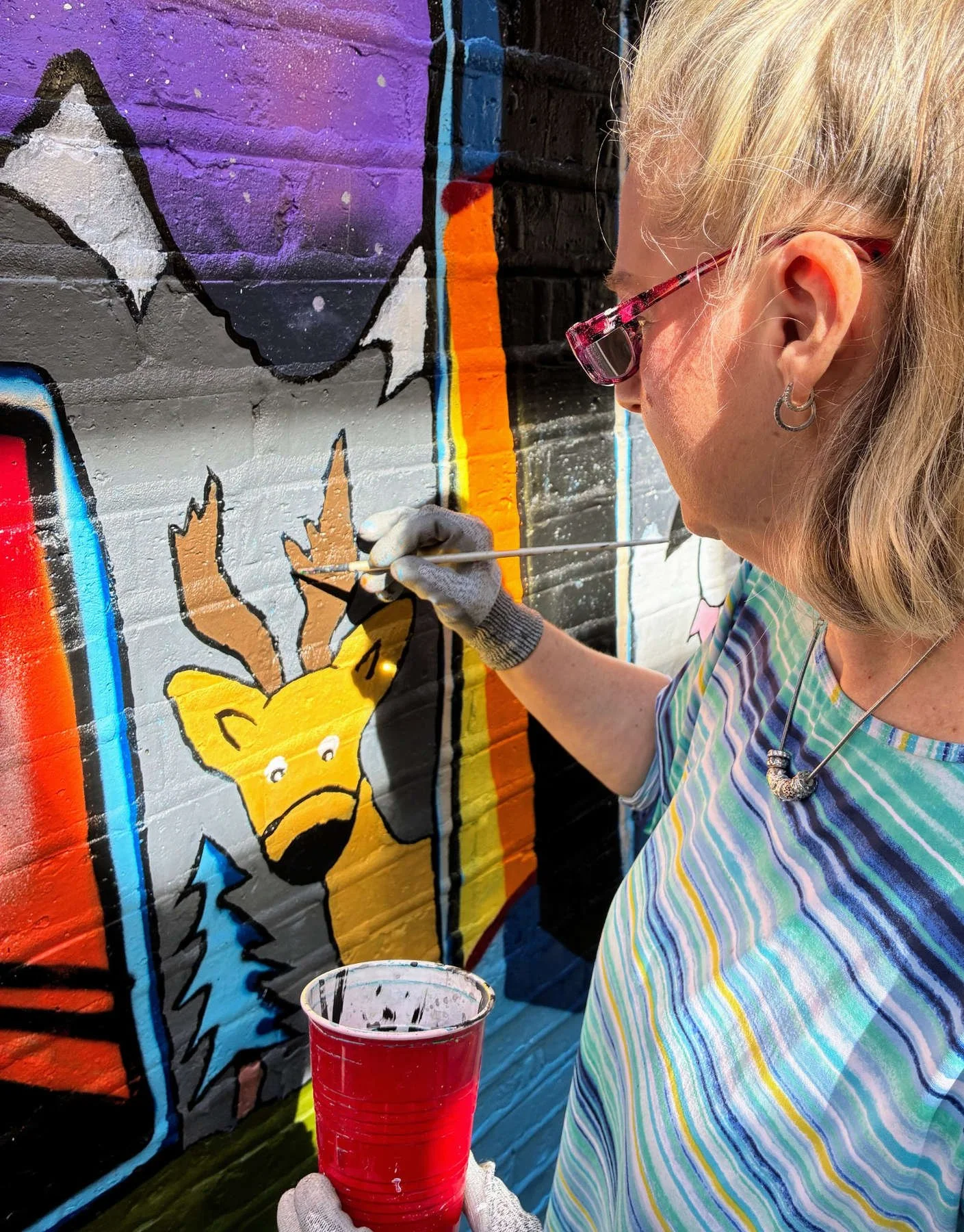 Community volunteer using a paintbrush to add details to a deer illustration within the colorful Lincoln Square community mural on a blue brick wall in Chicago.