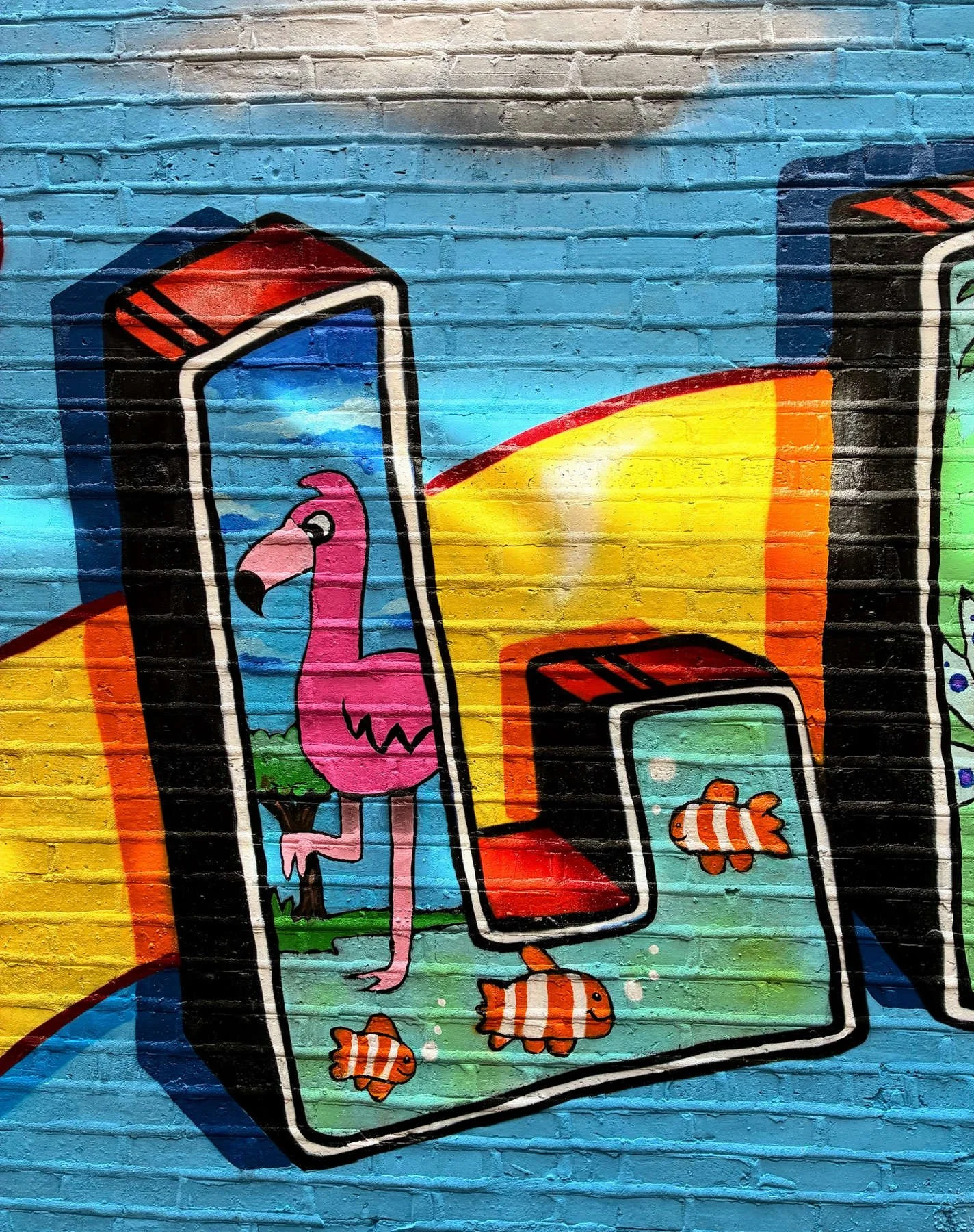 Close-up of the Lincoln Square community mural featuring a letter "L" with pink flamingo and orange clownfish within colorful three-dimensional graffiti lettering on a bright blue brick wall.