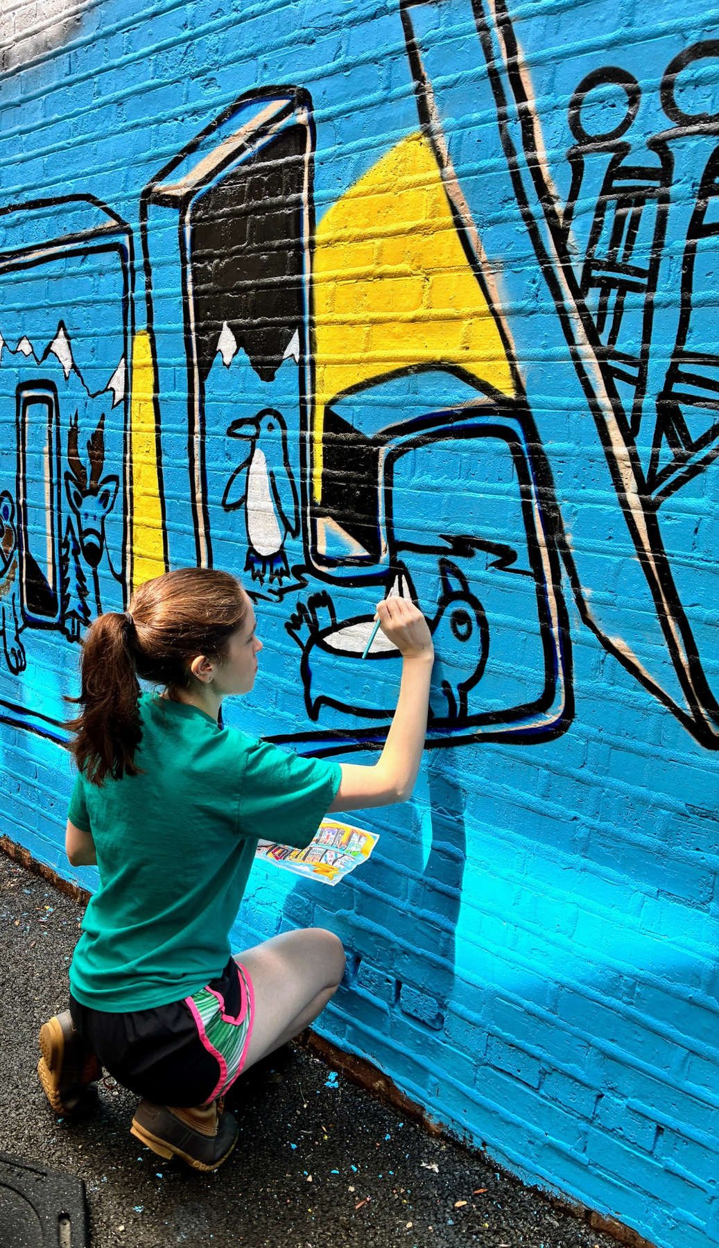 Community volunteer using a paintbrush to add colorful animal illustrations within the graffiti-style lettering of the Lincoln Square Mural Project 2026 in Chicago.