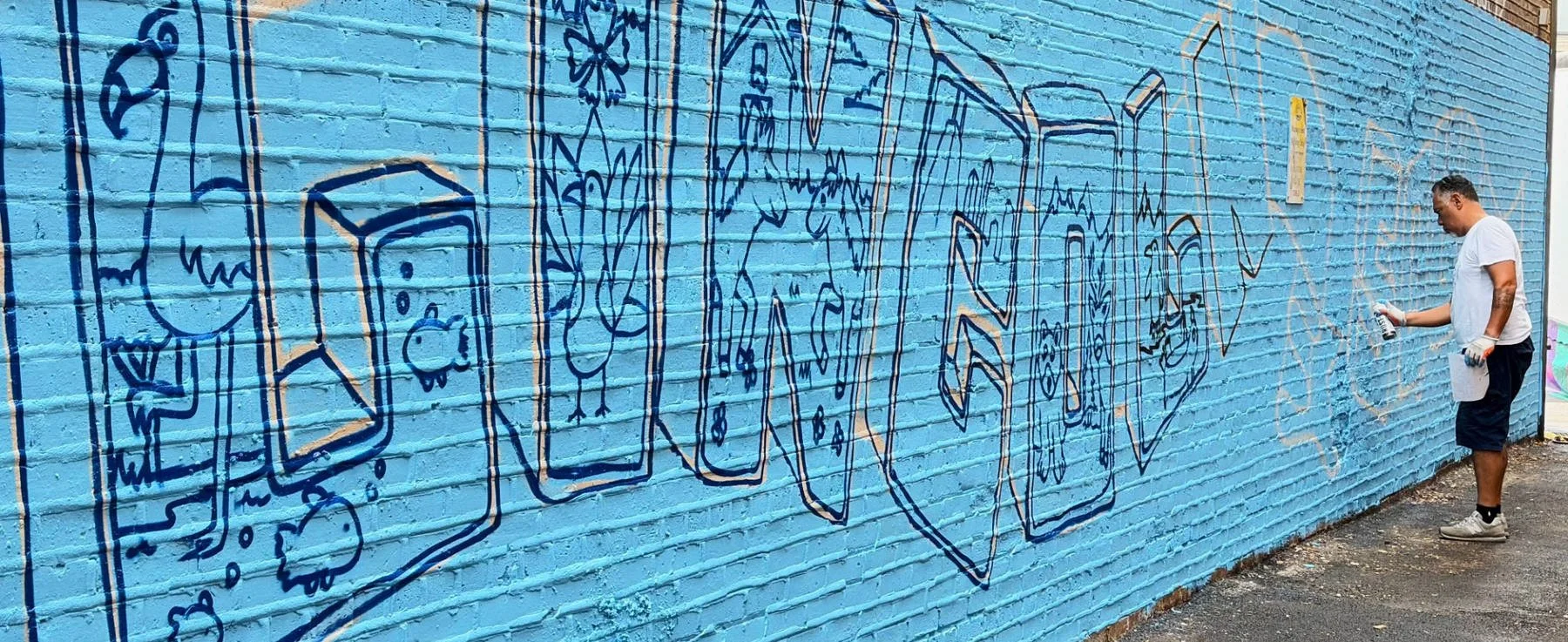 Nino “Tsel One/Tsel1” Rodriguez, lead artist of the Lincoln Square Mural Project 2026, outlining the graffiti-style lettering and animal illustrations on a blue brick wall in preparation for community painting.