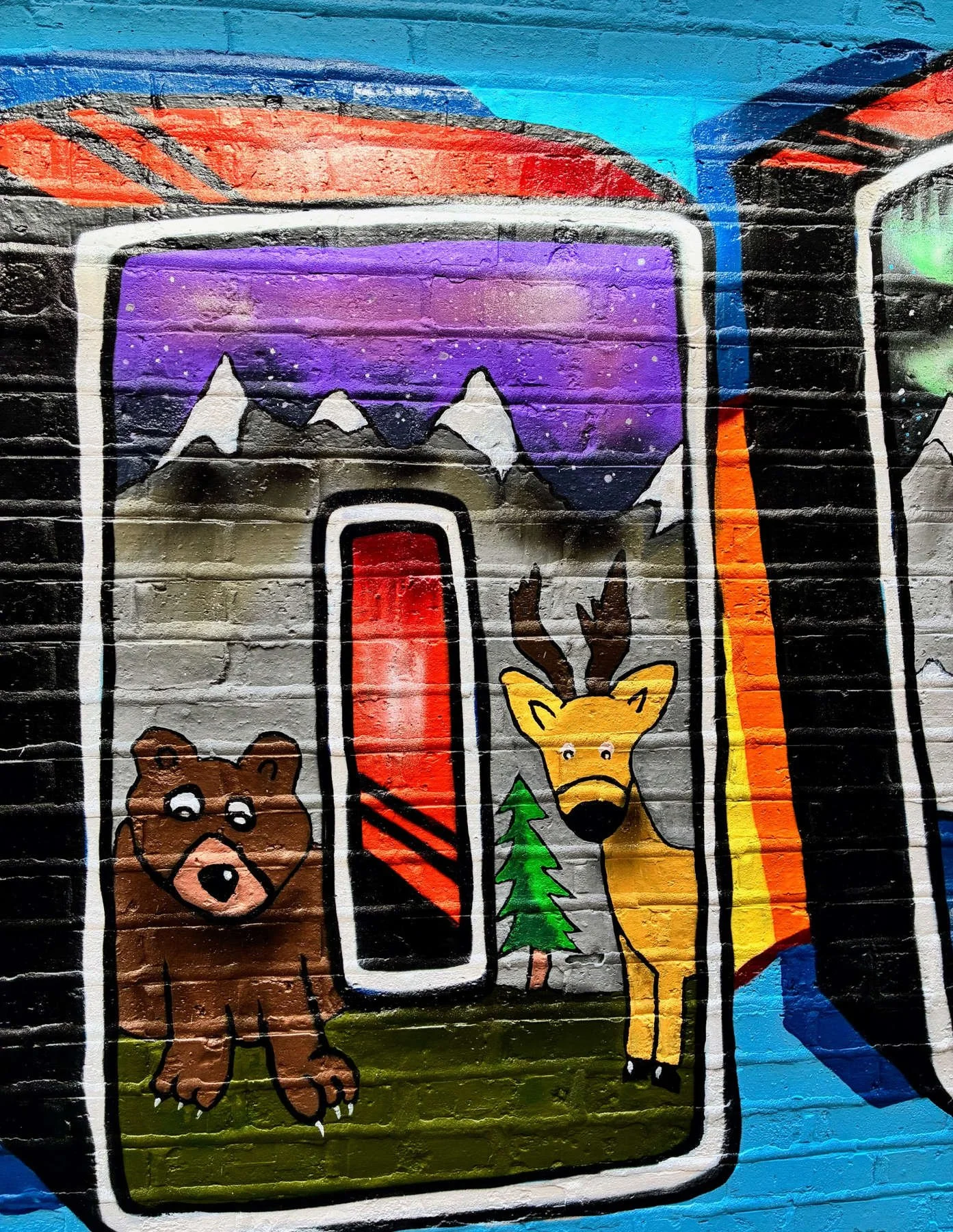 Close-up of the Lincoln Square mural featuring a letter "O" with brown bear, yellow deer, evergreen tree, and snow-capped mountains within colorful graffiti-style lettering on a blue brick wall.