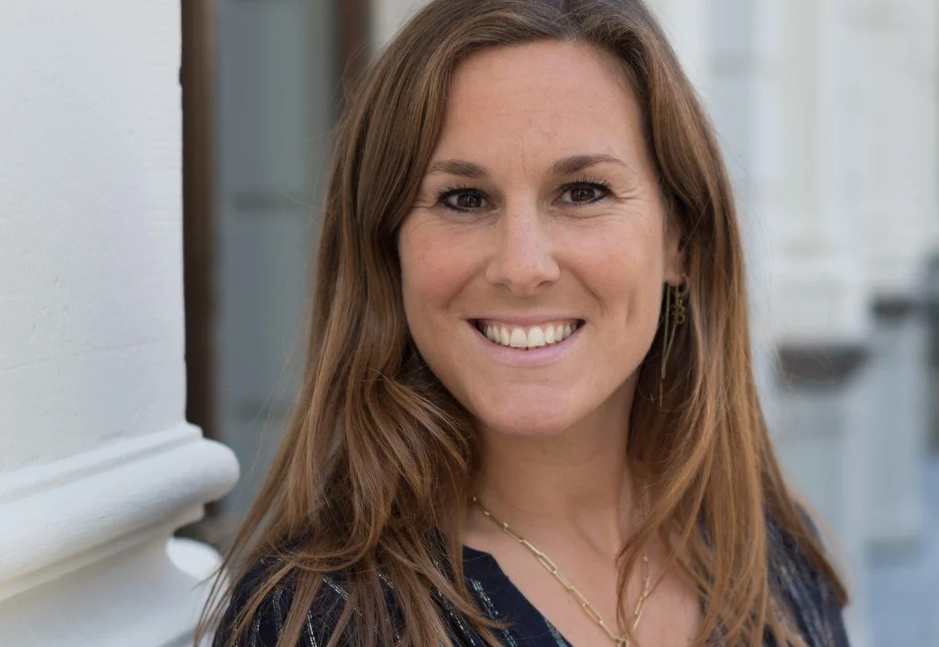 A woman with brown hair smiling outdoors, with a blurred background and white walls.