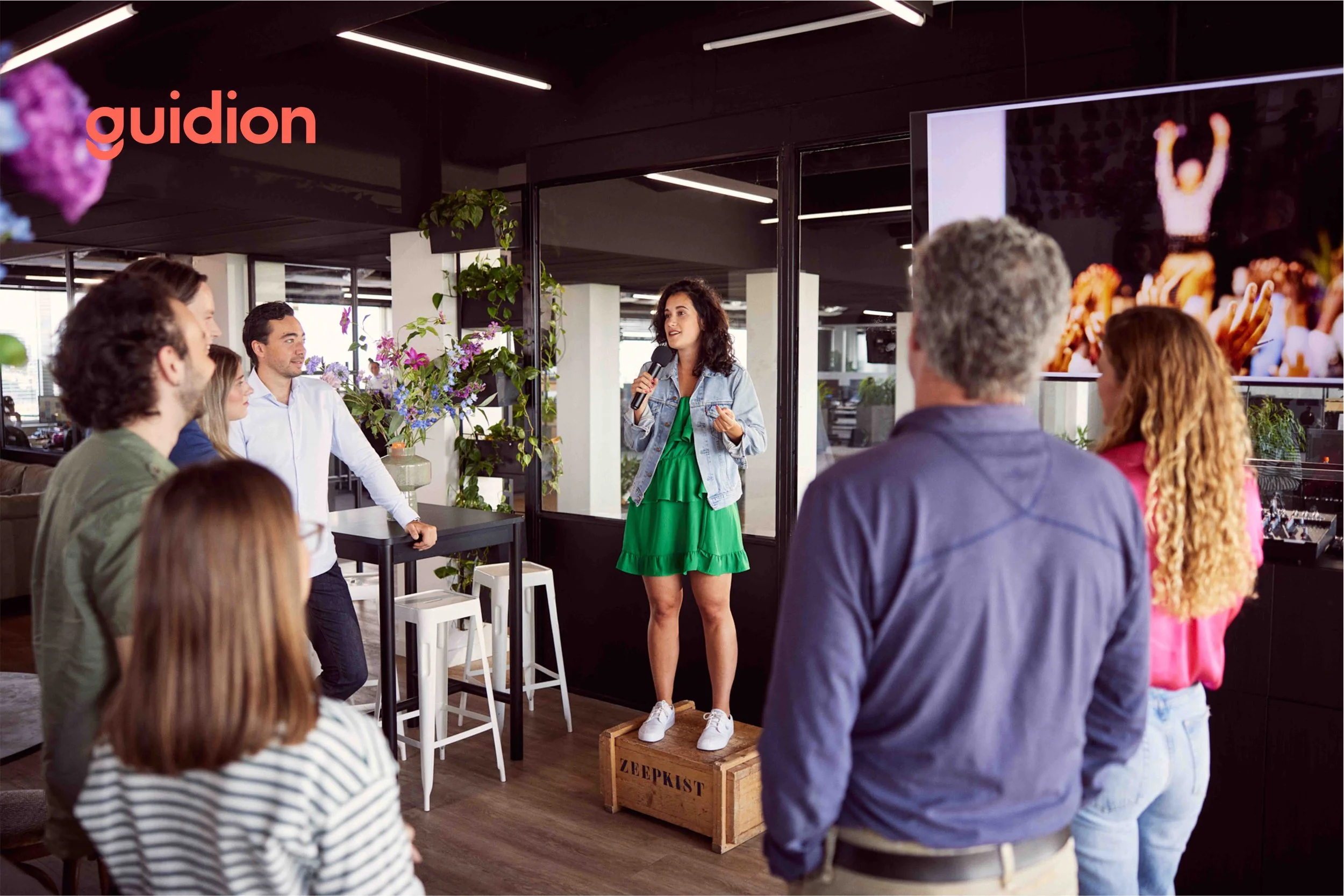 A woman in a green dress and denim jacket stands on a wooden crate and speaks into a microphone to a small group of seated and standing people in an indoor space decorated with plants and flowers during a presentation or performance.