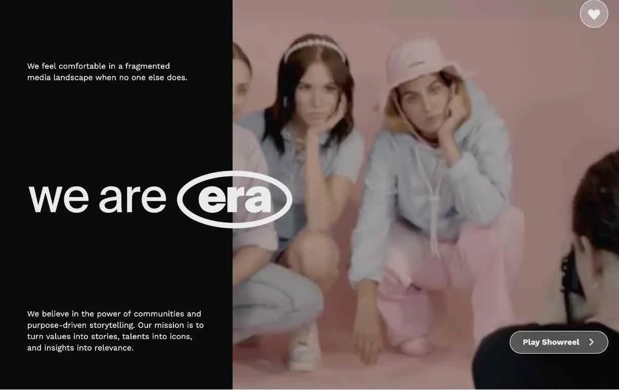 Group of three young women sitting on the floor against a pink background, with a black and white text overlay on the left side. The women are wearing casual clothes, one with a white hoodie and pink pants, another with a gray hoodie and gray shorts, and the third with a blue shirt and pink shorts. One is wearing a pink hat and has headphones around her neck. A photographer is seen in the foreground taking their picture.