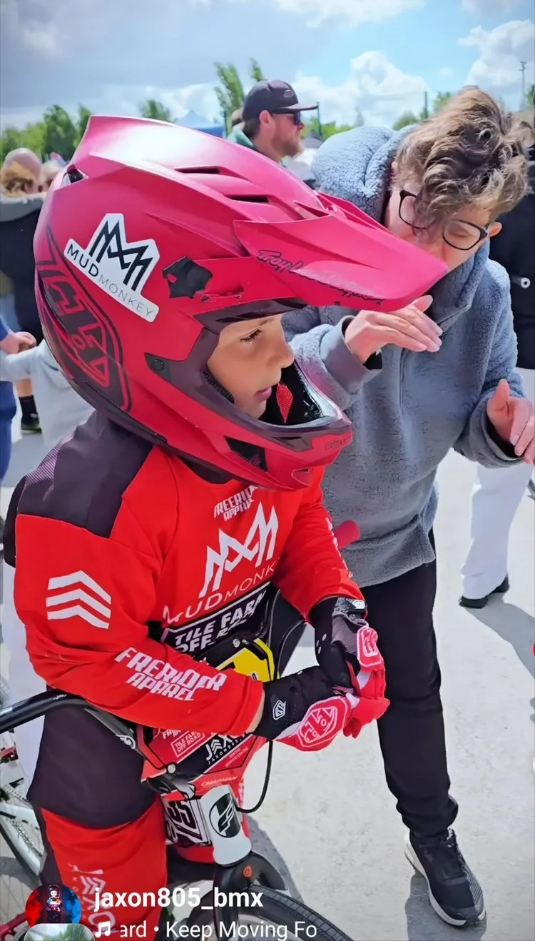 A young BMX rider wearing a red helmet, red jersey, and gloves standing next to a woman with glasses and curly hair, outdoors on a cloudy day surrounded by other people.