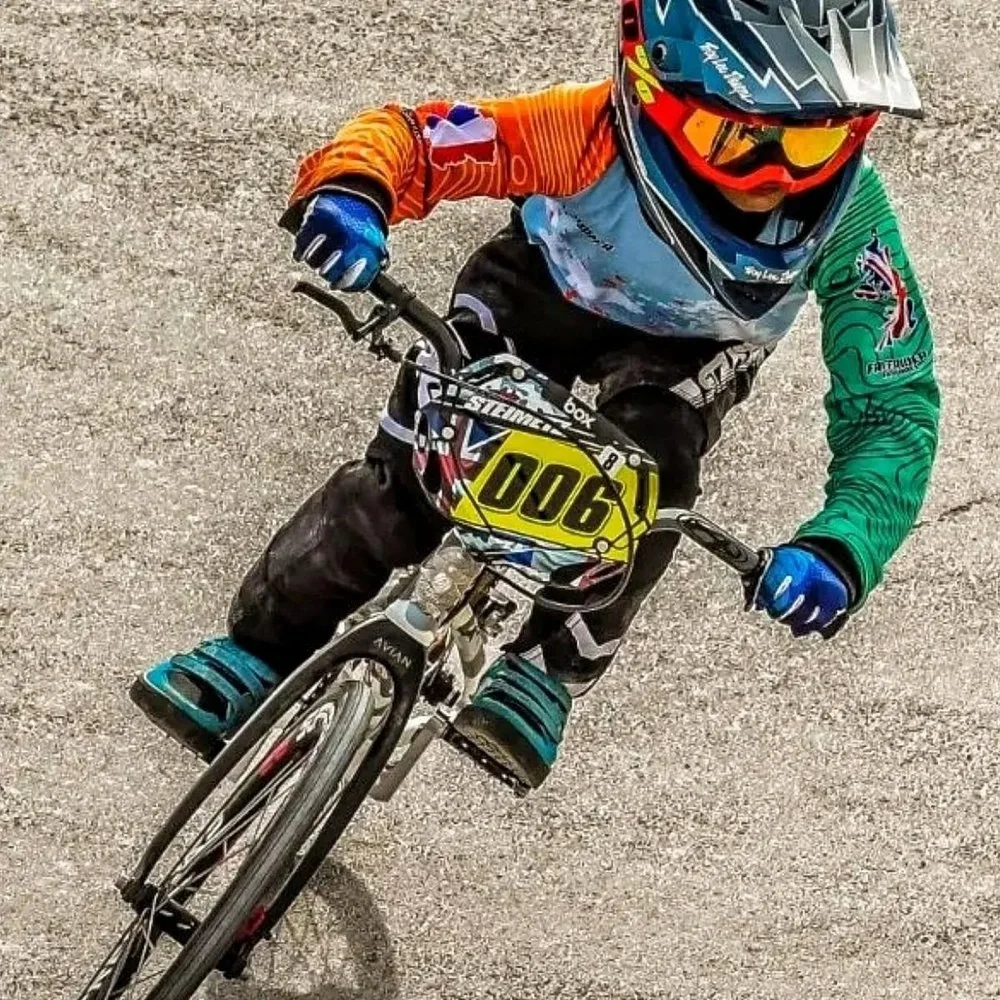 A young person riding a BMX bike on a gravel surface, wearing a helmet, goggles, and colorful protective gear, including gloves and padded clothing.