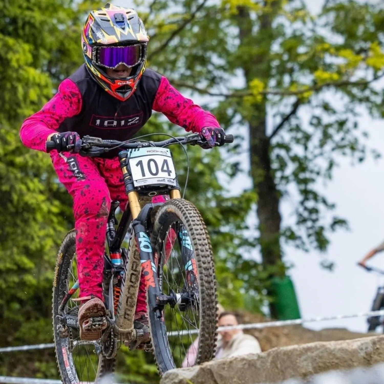 A mountain biker wearing a pink and black custom designed jersey and pants, helmet, and goggles rides over a rocky trail with greenery in the background.