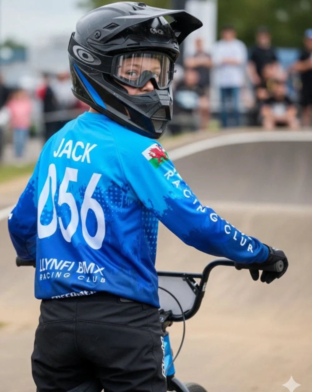 A young person wearing a black helmet and goggles, blue jersey with the name Jack and number 056, holding onto a BMX bike, with a crowd of spectators in the background at a BMX racing event.